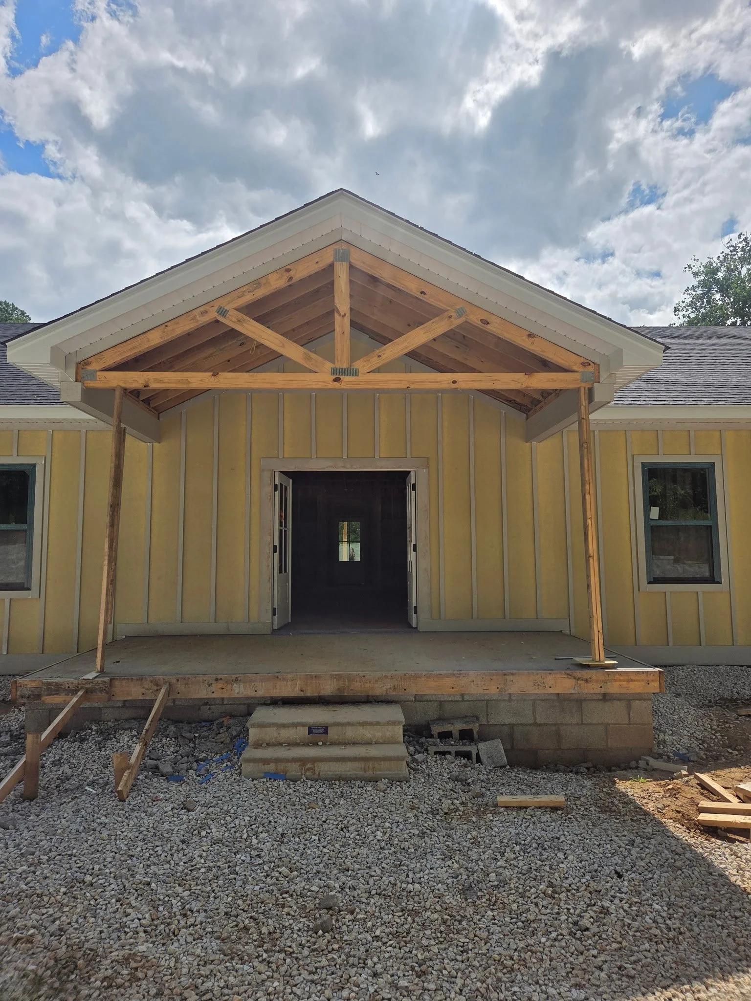 The image shows a house under construction, with wooden framing and support beams in place for a new porch or entryway, with a gravel ground and a cloudy sky overhead.