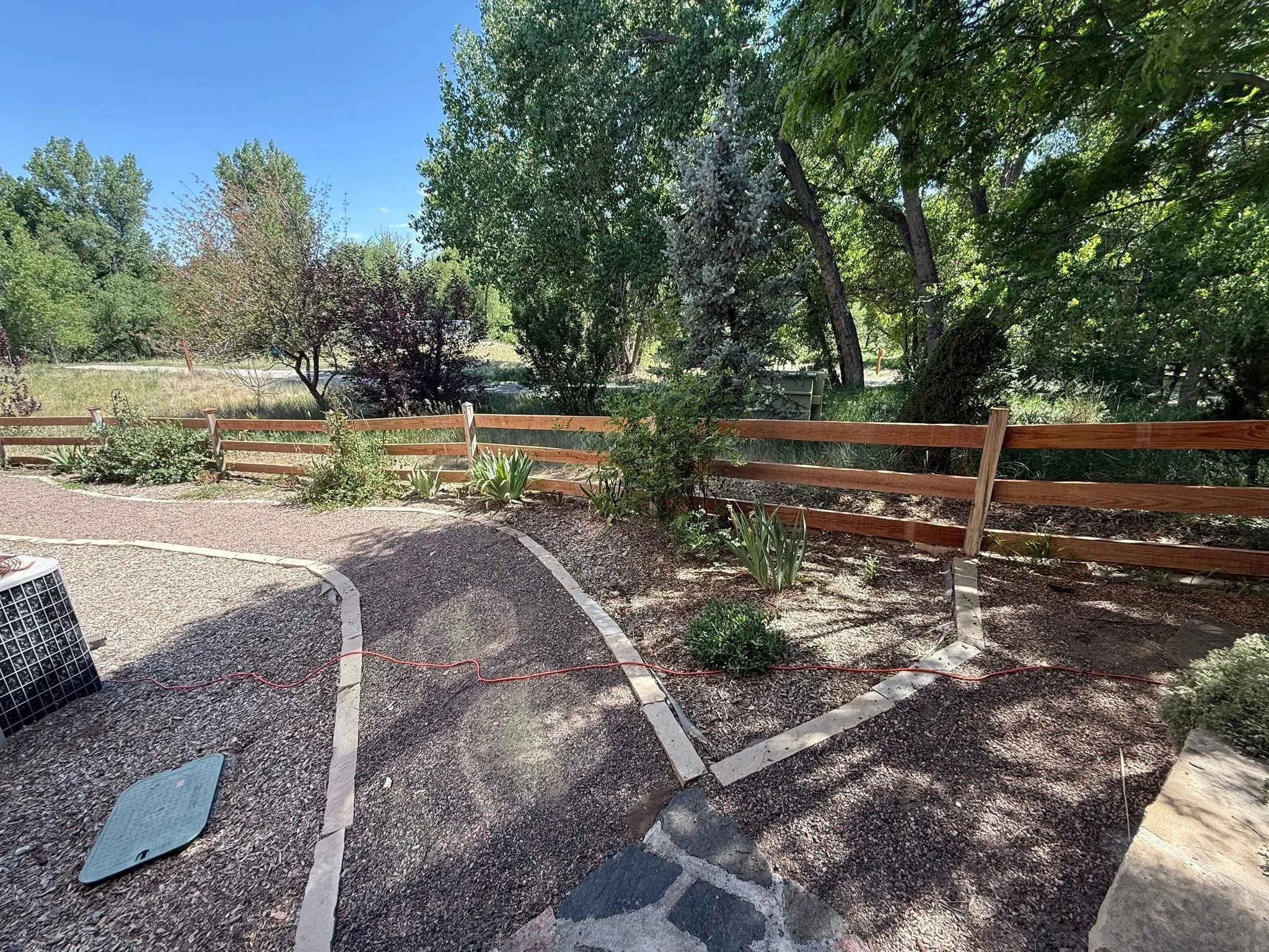 A landscaped backyard with trees, shrubs, a wooden fence, and a gravel pathway, with part of solar panels and outdoor equipment visible in the foreground.