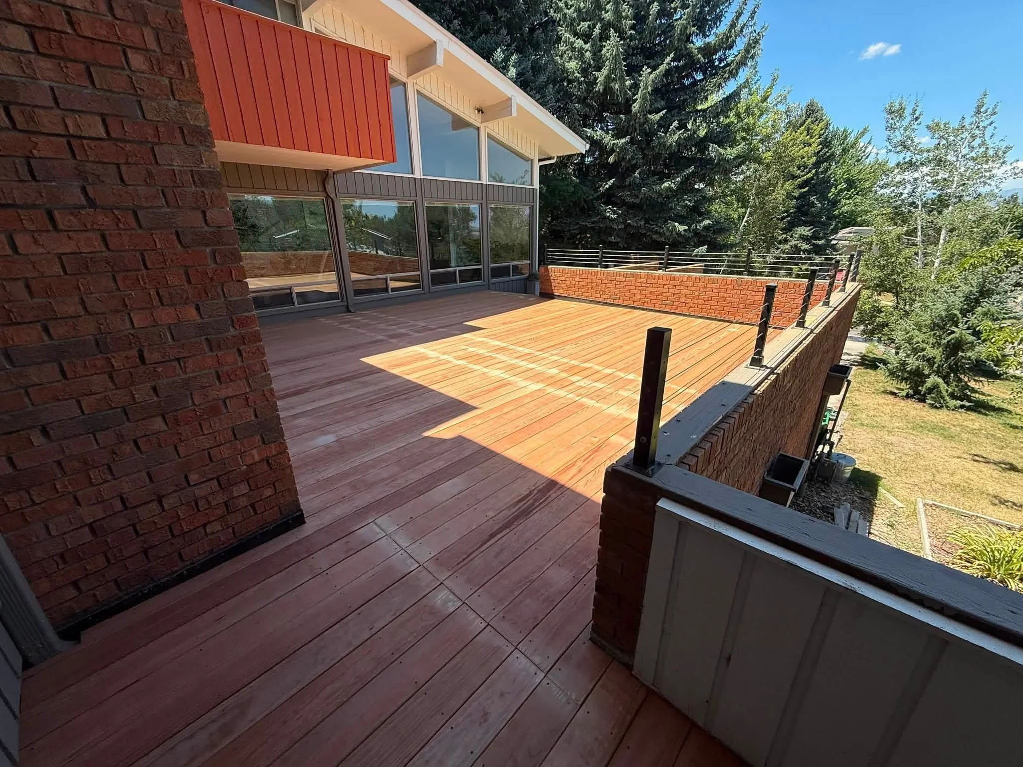 Wooden balcony with brick walls and black metal railing, overlooking a yard with green trees on a sunny day.