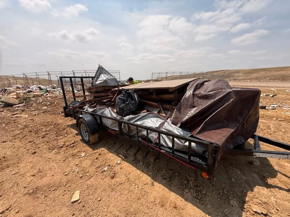 Trailer loaded with miscellaneous materials and covered with tarps on a dirt road in an open landscape with a fence in the background.