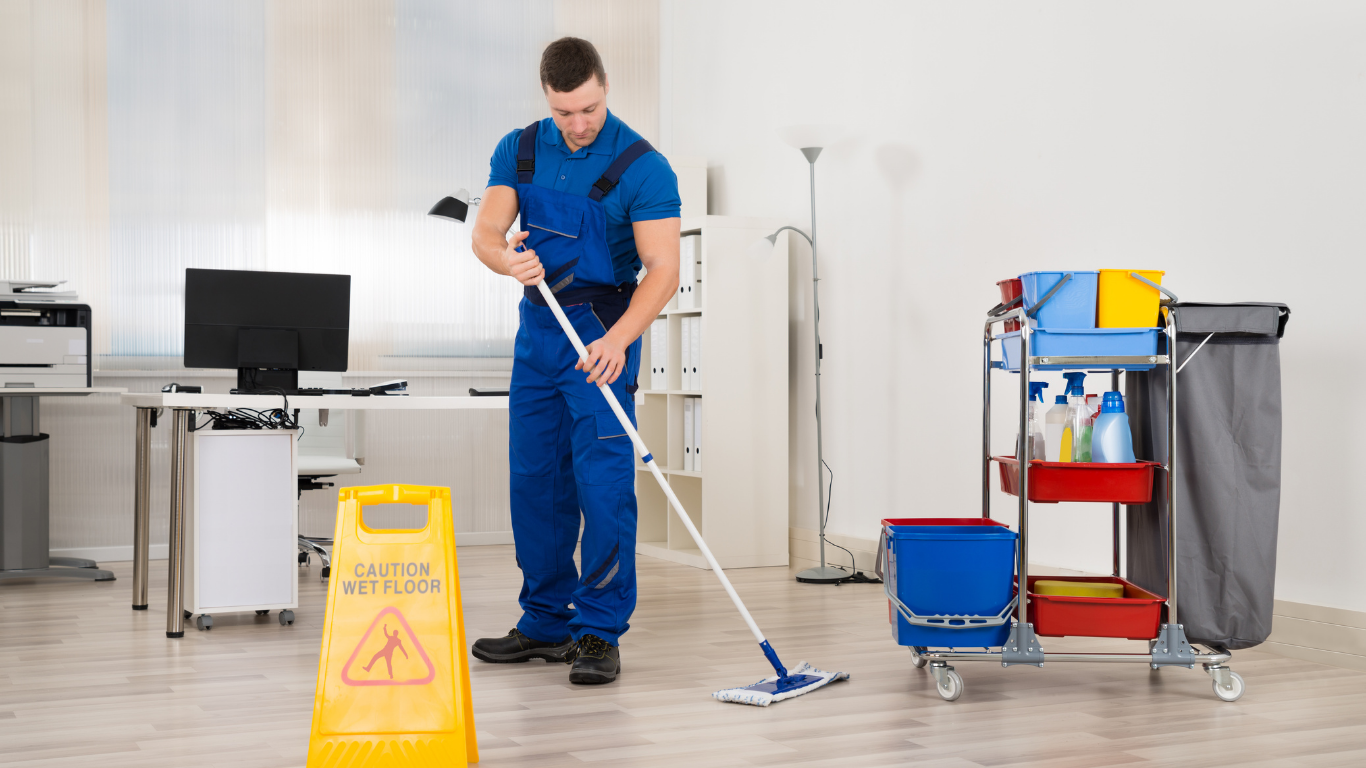 A janitor mopping the floor in an office with cleaning supplies on a cart and a yellow caution wet floor sign.