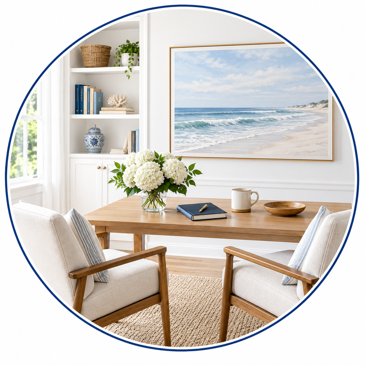A cozy dining area with a wooden table, two armchairs with white cushions, and a vase of white flowers. Behind the table, a large framed picture of a beach scene hangs on the wall. To the left, a built-in white shelf holds books and decorative items, with a window allowing natural light to illuminate the room.