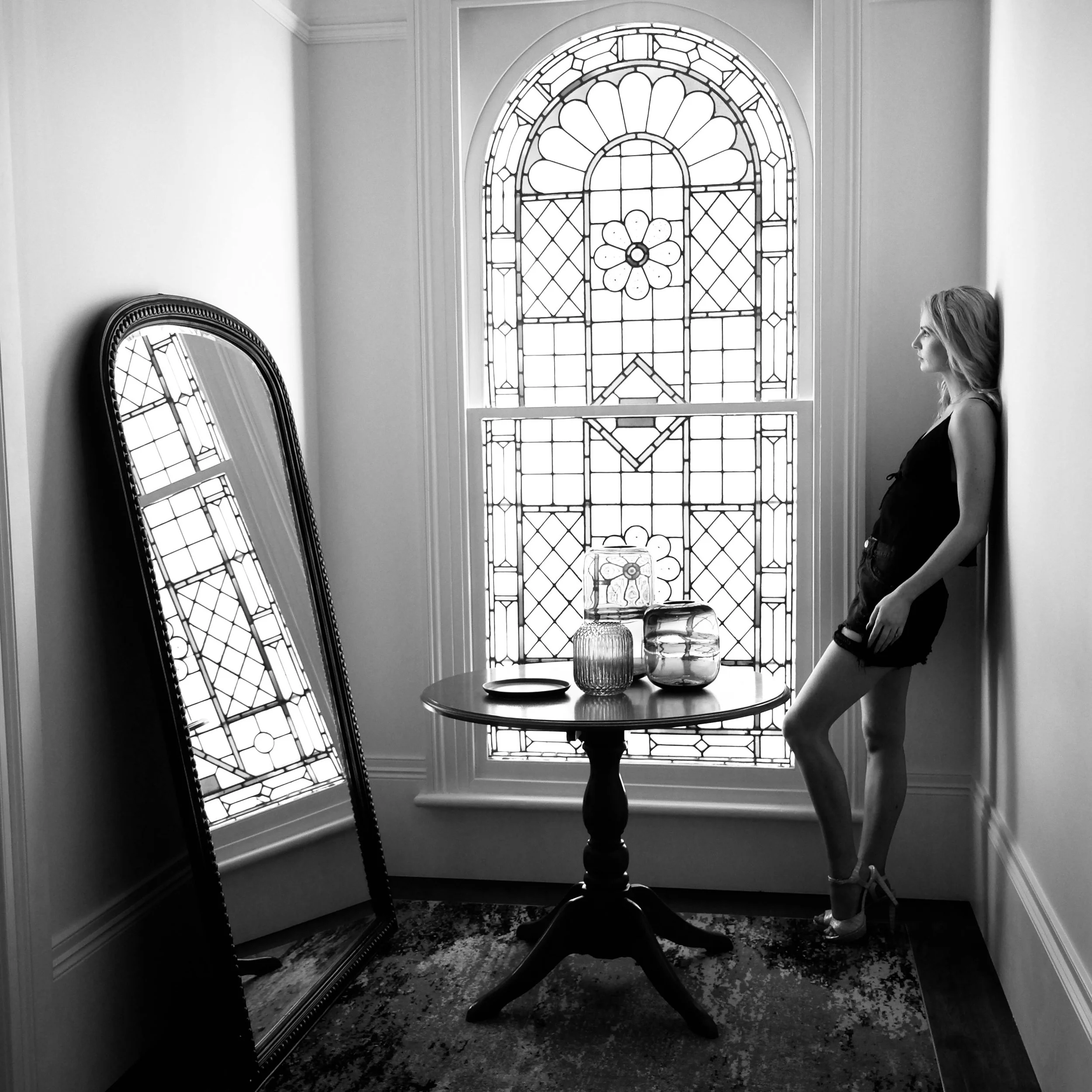 A woman standing in front of a large decorative stained glass window with a floral design, with a tall oval mirror reflecting a portion of the window and a table with glass and ceramic vases, in a room with a patterned rug.