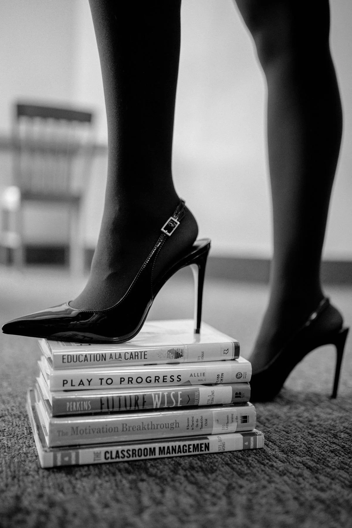 Black high-heeled shoe with a zipper on the ankle, placed on top of a stack of books. The background shows a chair and a bottle on a table. The image is in black and white.