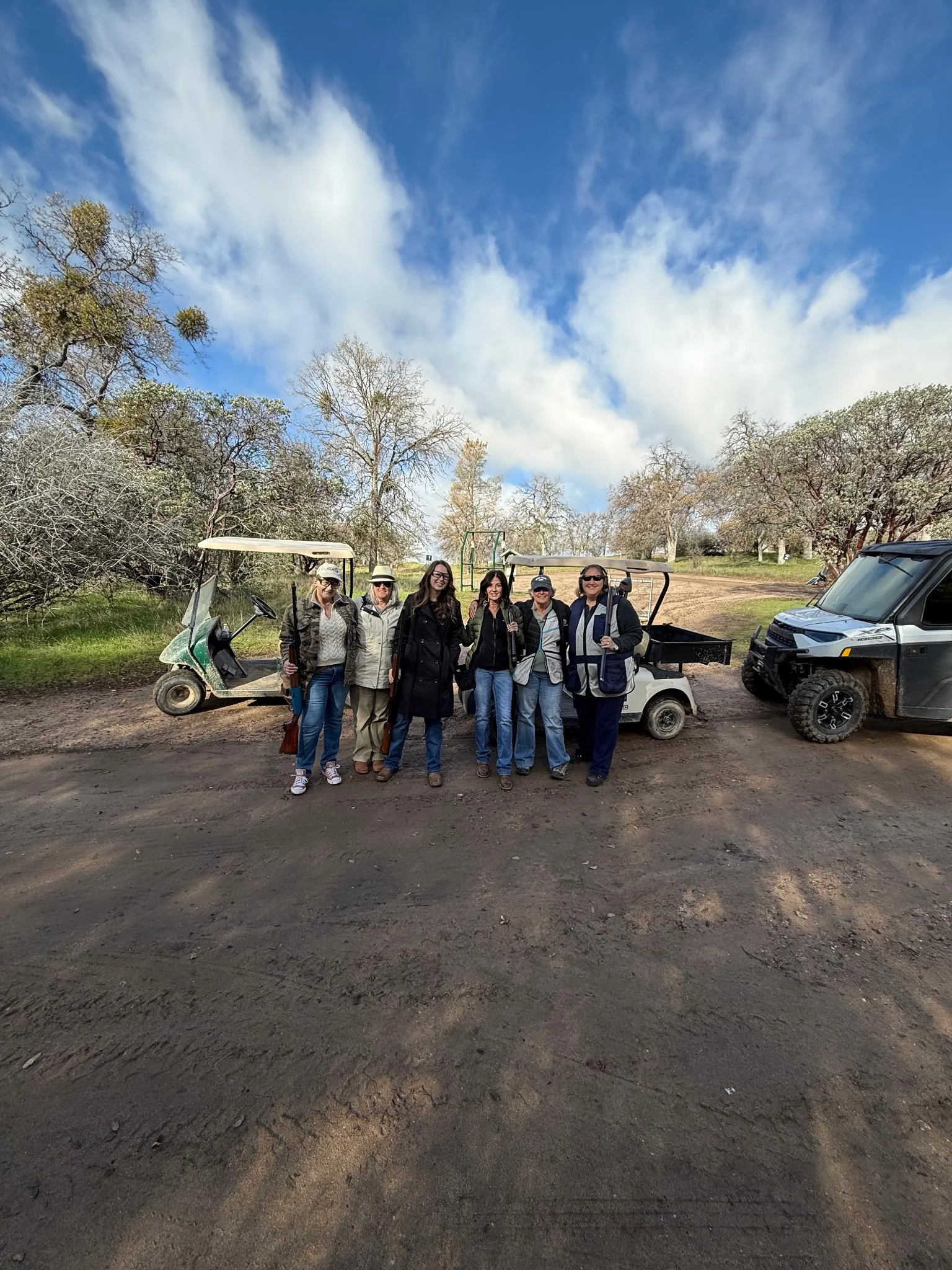 A group of six people standing together outdoors beside a golf cart and a utility vehicle, with trees and a partly cloudy sky in the background.