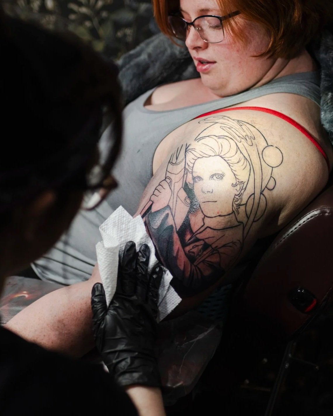 A woman with glasses is getting a tattoo on her upper arm of a woman holding a lightsaber with a cosmic background.