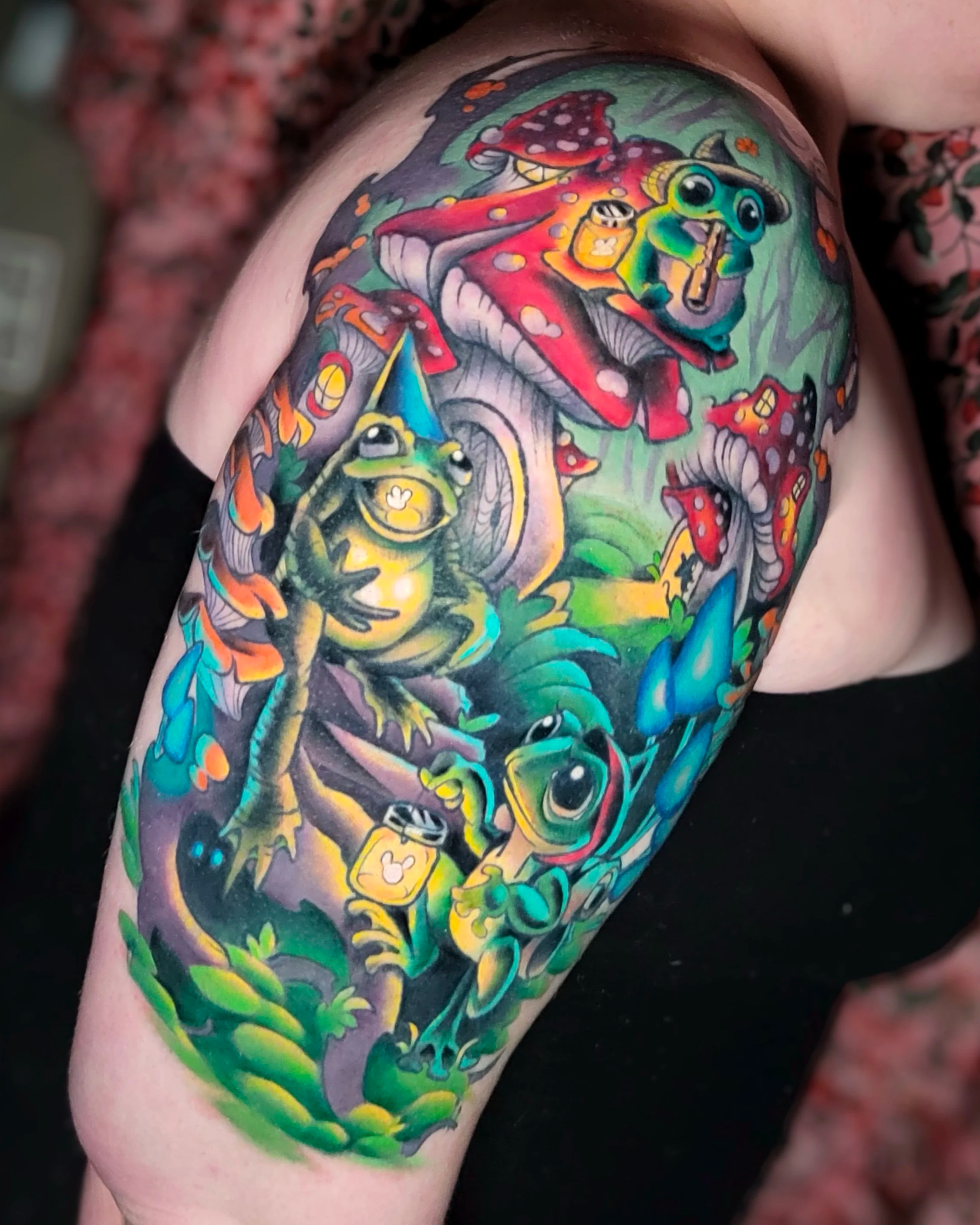 Colorful tattoo featuring cartoon-style frogs, mushrooms, and foliage on a person's upper arm.