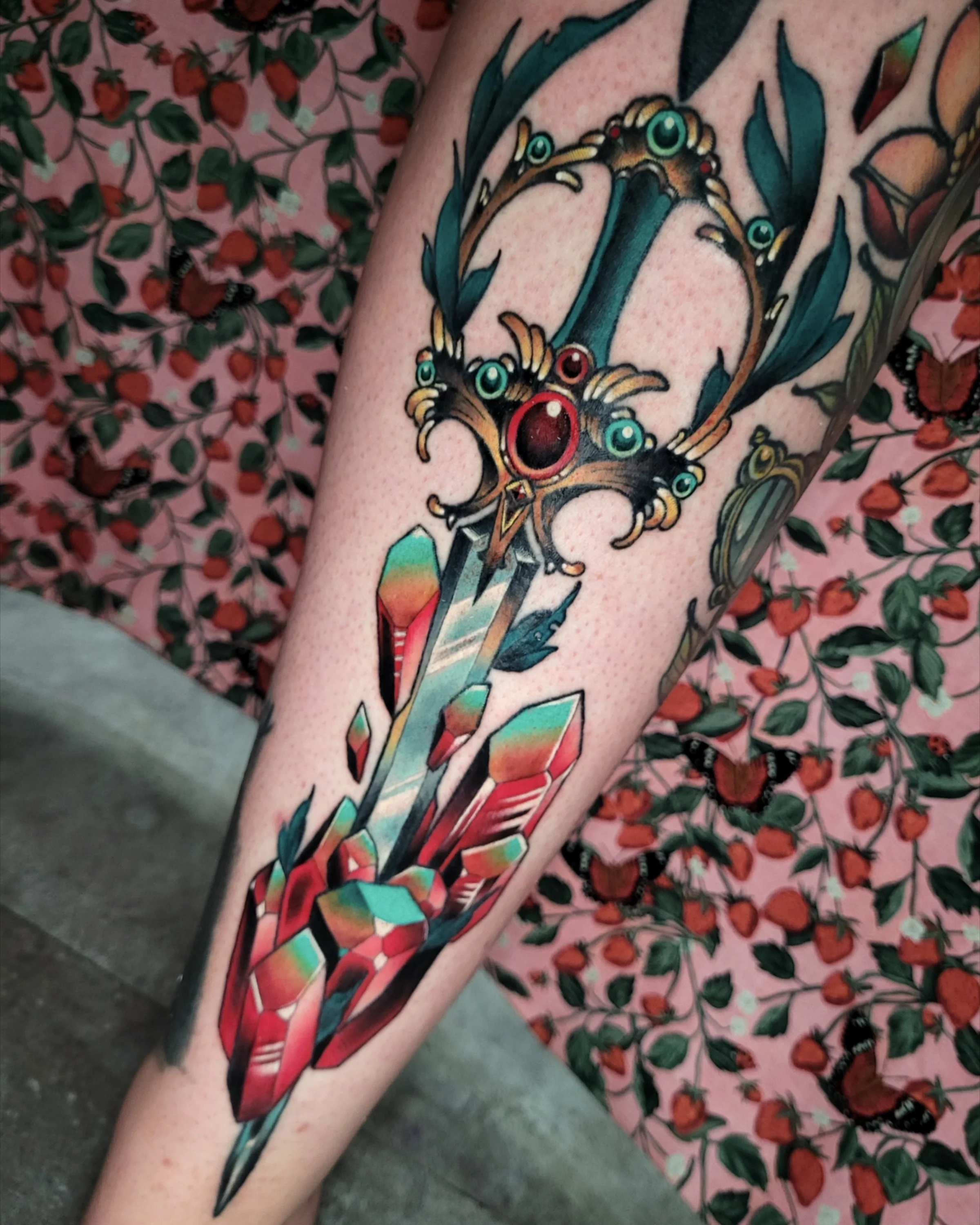 A colorful tattoo of a sword with gemstones, feathers, and decorative elements on a person's arm.
