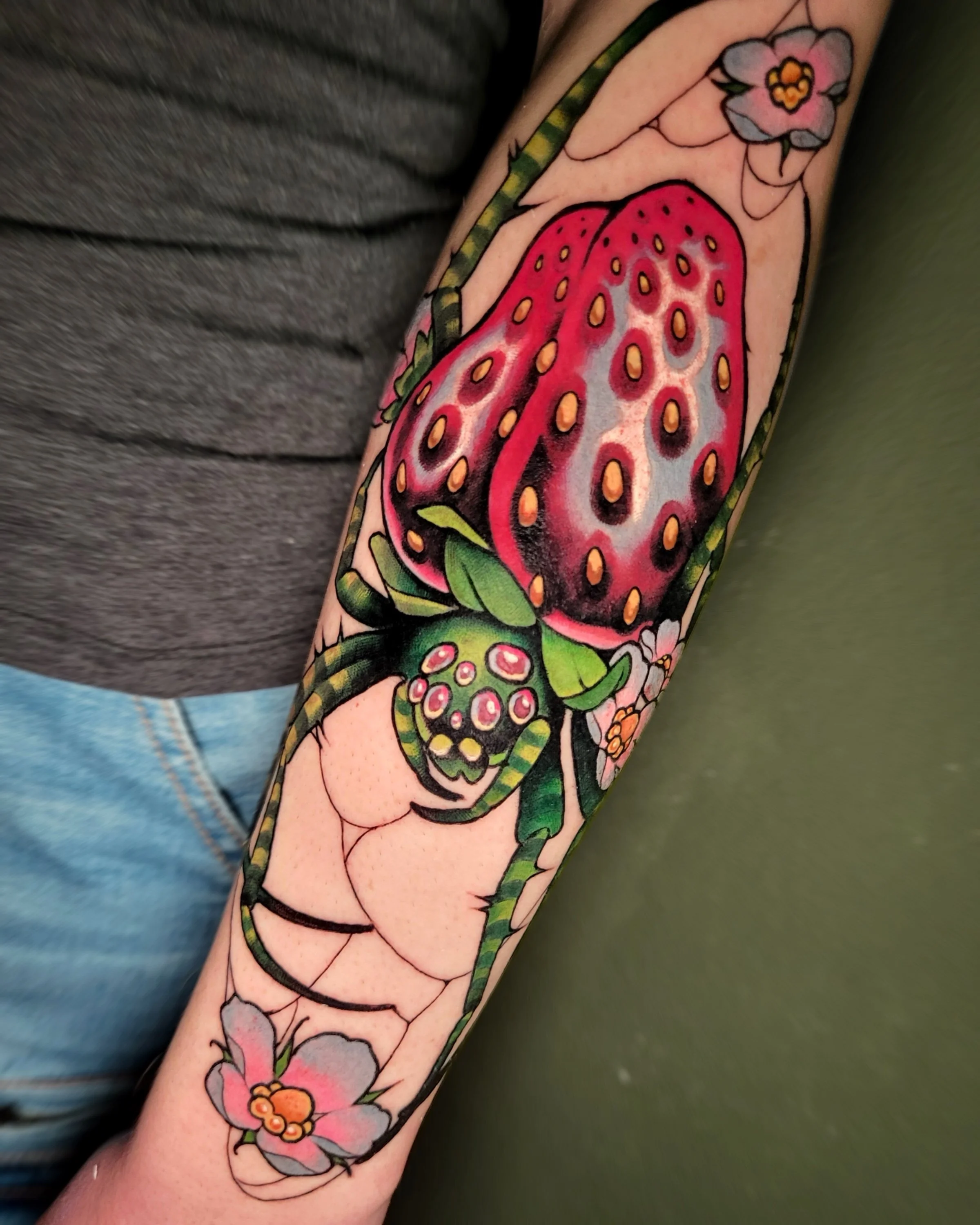 Colorful tattoo of a strawberry with a small pink flower and green leaves on a person's forearm.