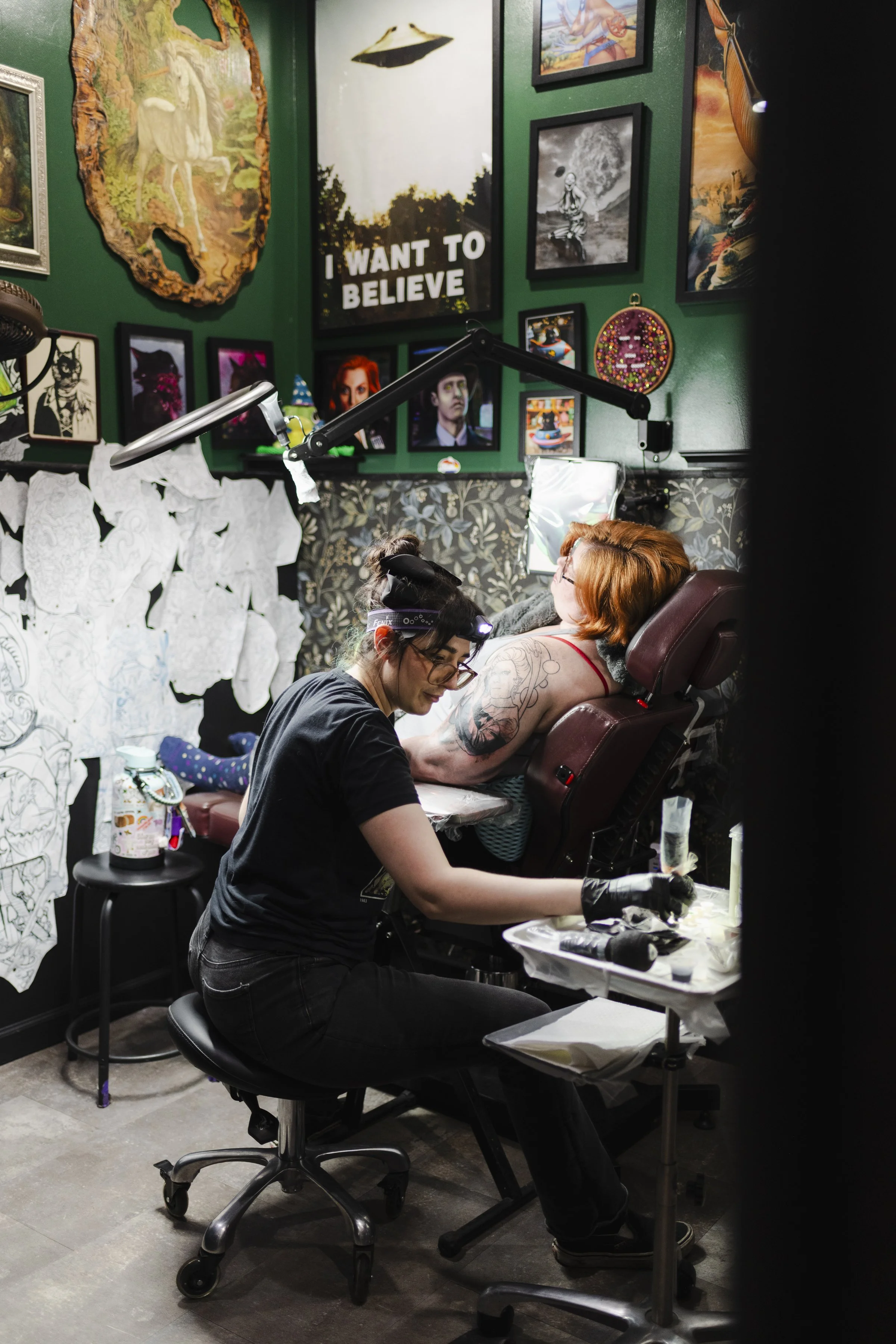 A person with red hair and tattooed arm lying on a chair, receiving a tattoo from a tattoo artist. The room has dark green walls decorated with various framed pictures and artwork, including a large