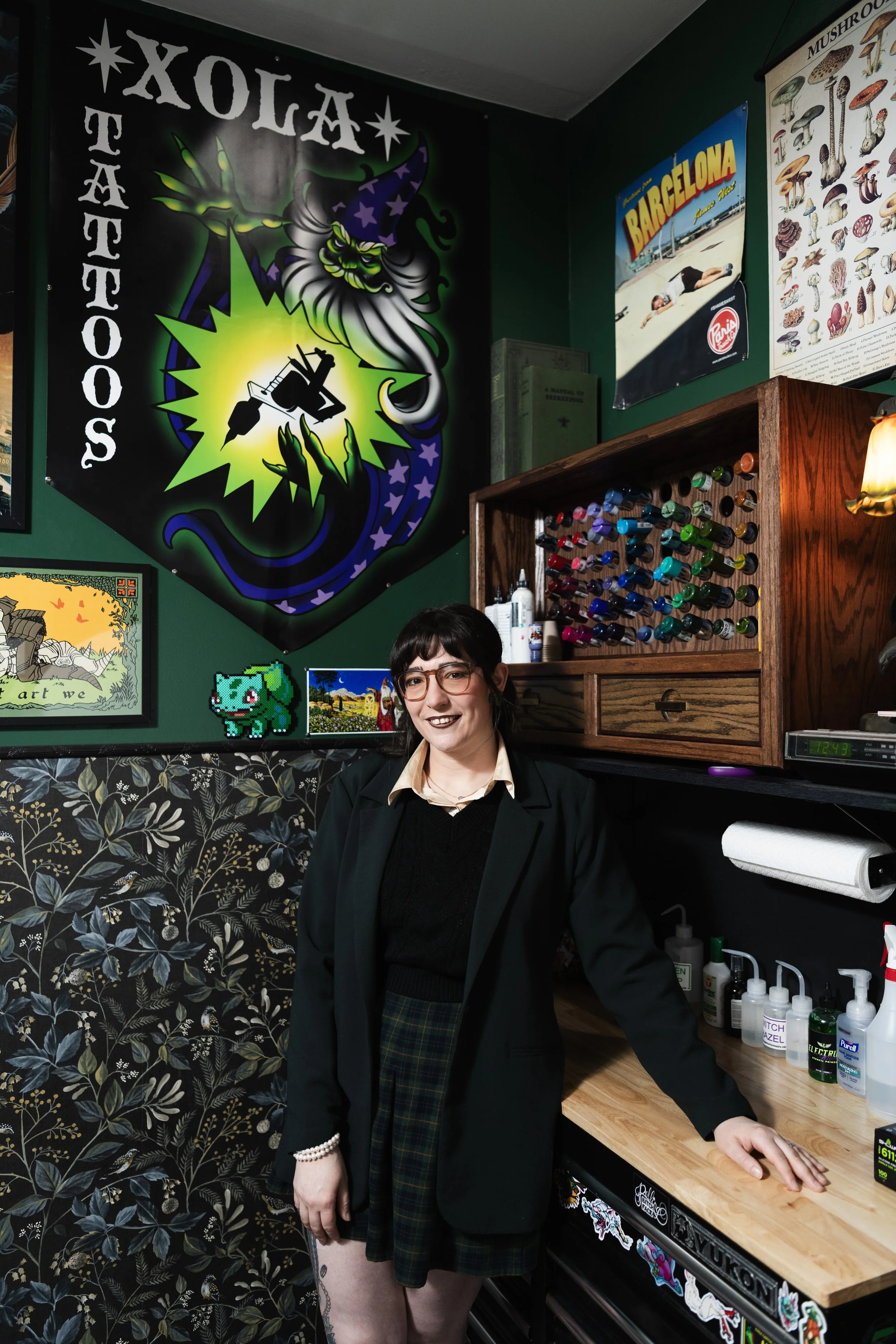 A woman with glasses, dark hair, and fair skin is smiling and standing behind a wooden counter in a room decorated with posters and artwork. She is wearing a black blazer, a cream-colored shirt, and plaid skirt. The background includes a large tattoo shop banner, a Barcelona travel poster, a detailed mushroom chart, and a pixel art character on the wall. The counter has various skincare and hygiene products, and a paper towel roll is visible. The wall features a floral patterned wallpaper.