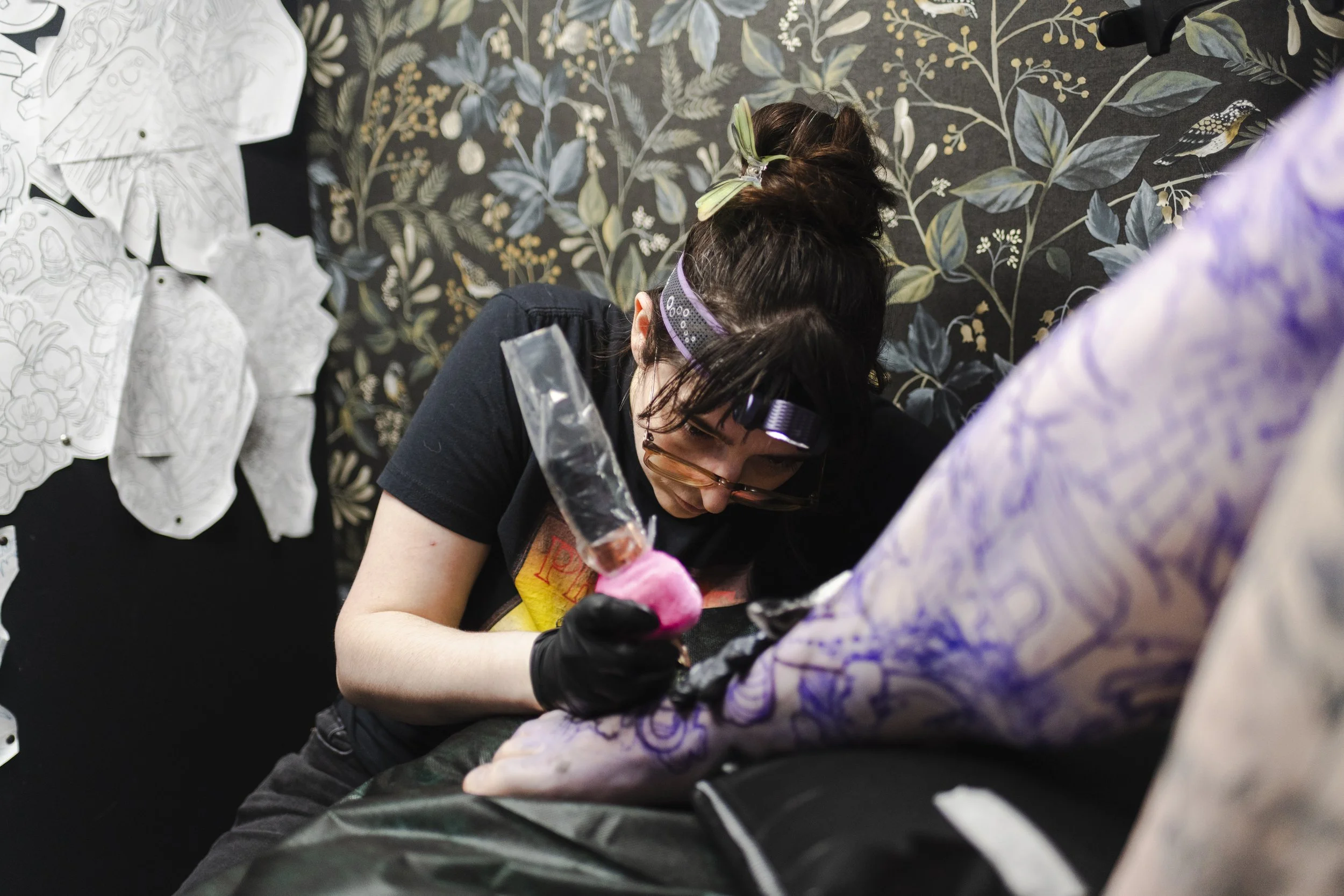 A person with glasses and a black shirt working on a craft project at a table covered with black fabric, surrounded by patterned paper and a purple-tinted fabric. A woman with brown hair, some with yellow streaks, wearing a gray headset, is leaning over, focused on her work. The background features a black wallpaper with a floral pattern with leaves and berries.