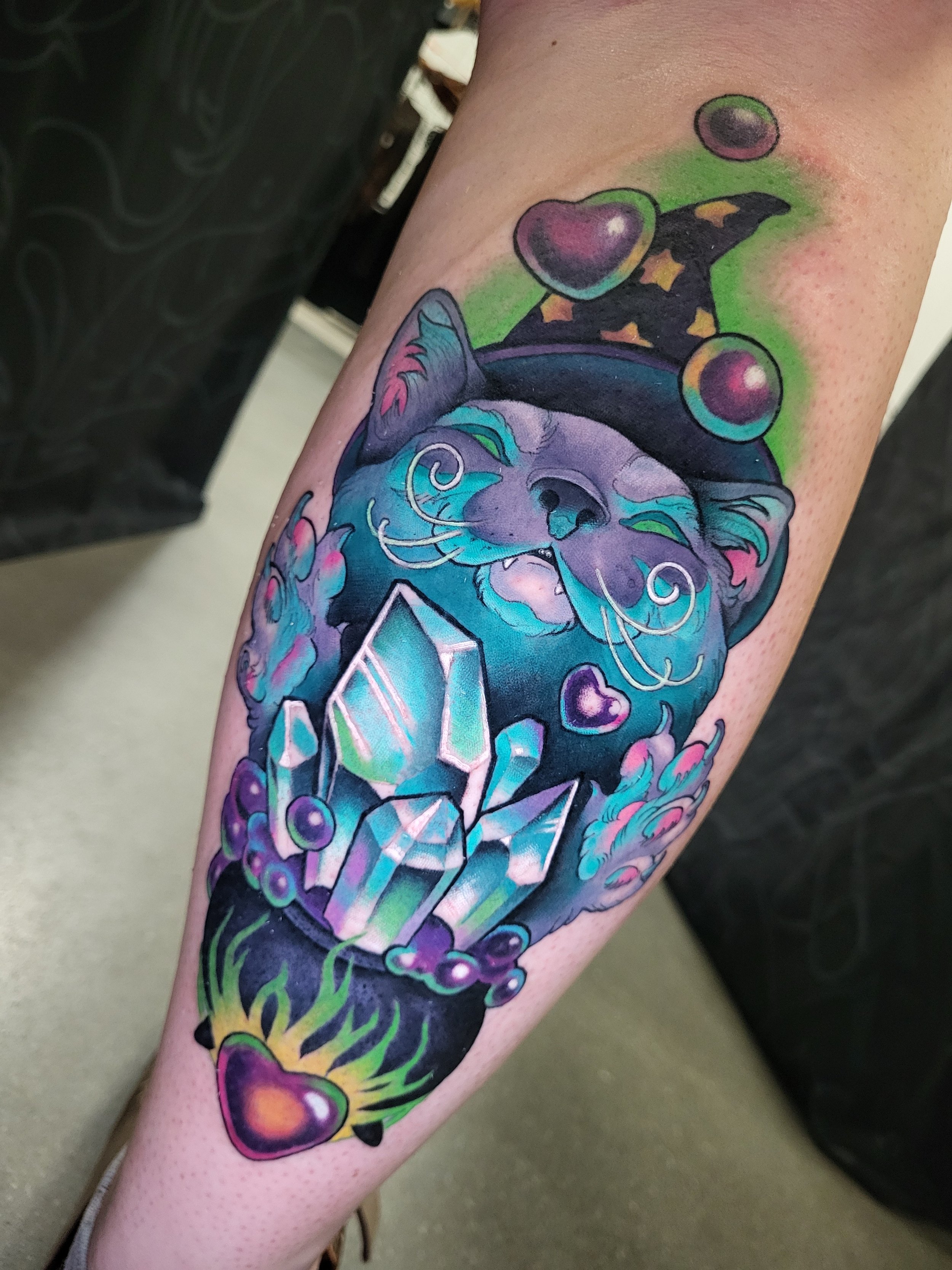 Colorful tattoo of a smiling cat wearing a wizard hat, surrounded by heart, star, and gemstone designs on a person's arm.