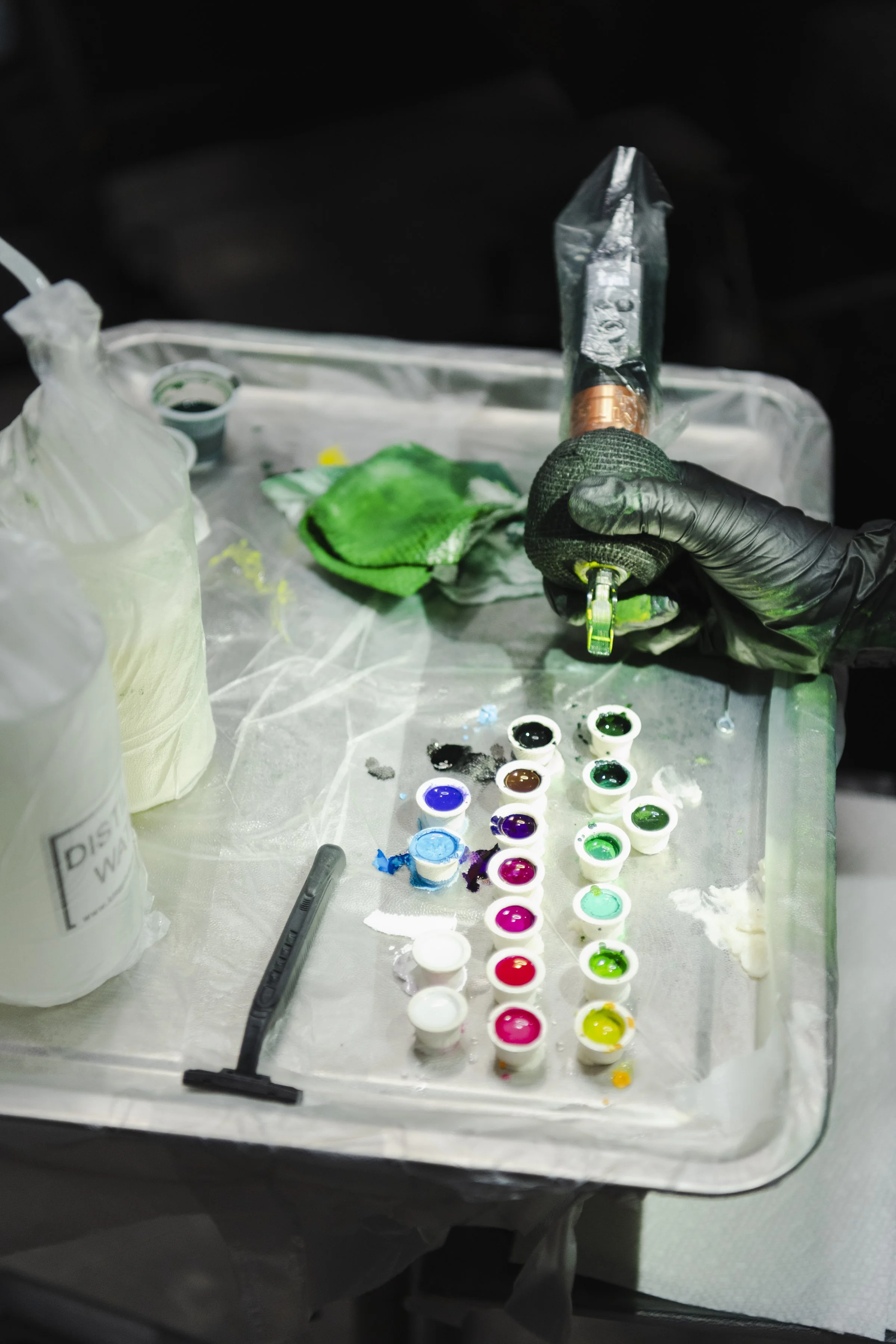A person wearing black gloves holds a silencer for a tattoo machine over a table covered with tattoo ink cups arranged in a rainbow pattern. The table has tattoo supplies including ink bottles, a green cloth, and various tattoo tools.