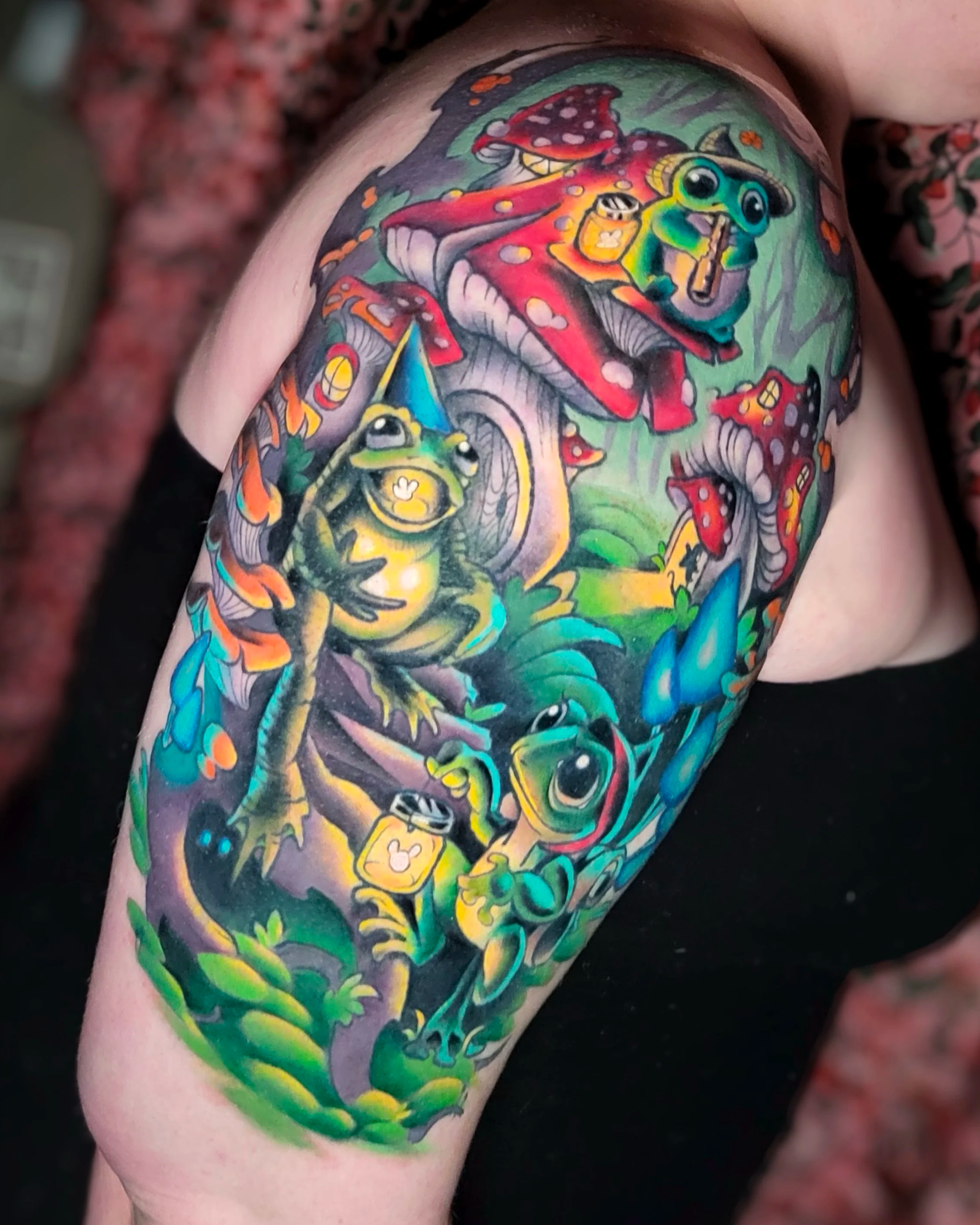 Colorful tattoo of frogs and mushrooms on person's upper arm with vibrant greens, reds, and yellows.