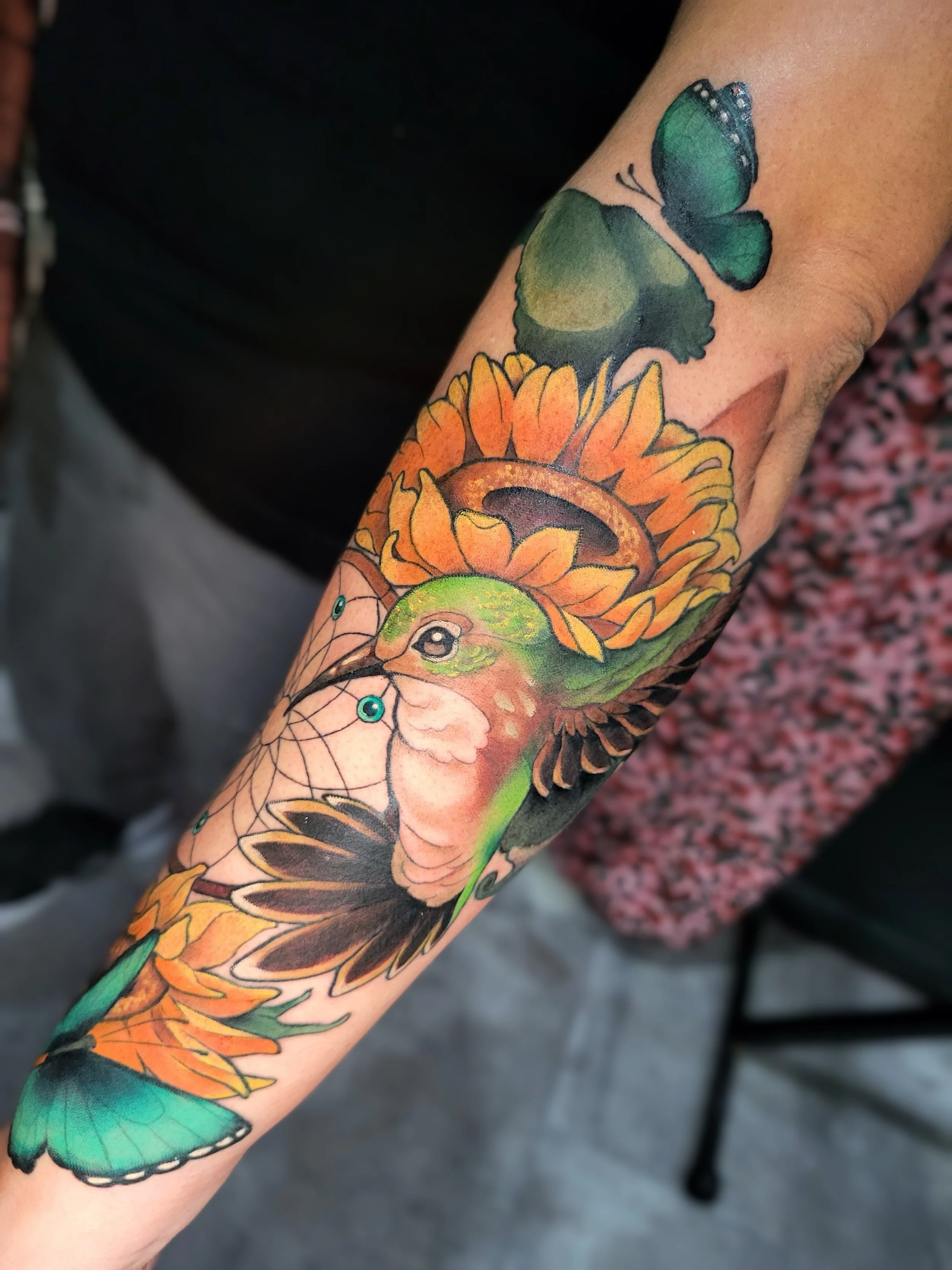 Colorful tattoo of a butterfly, sunflower, and bird on a person's arm.