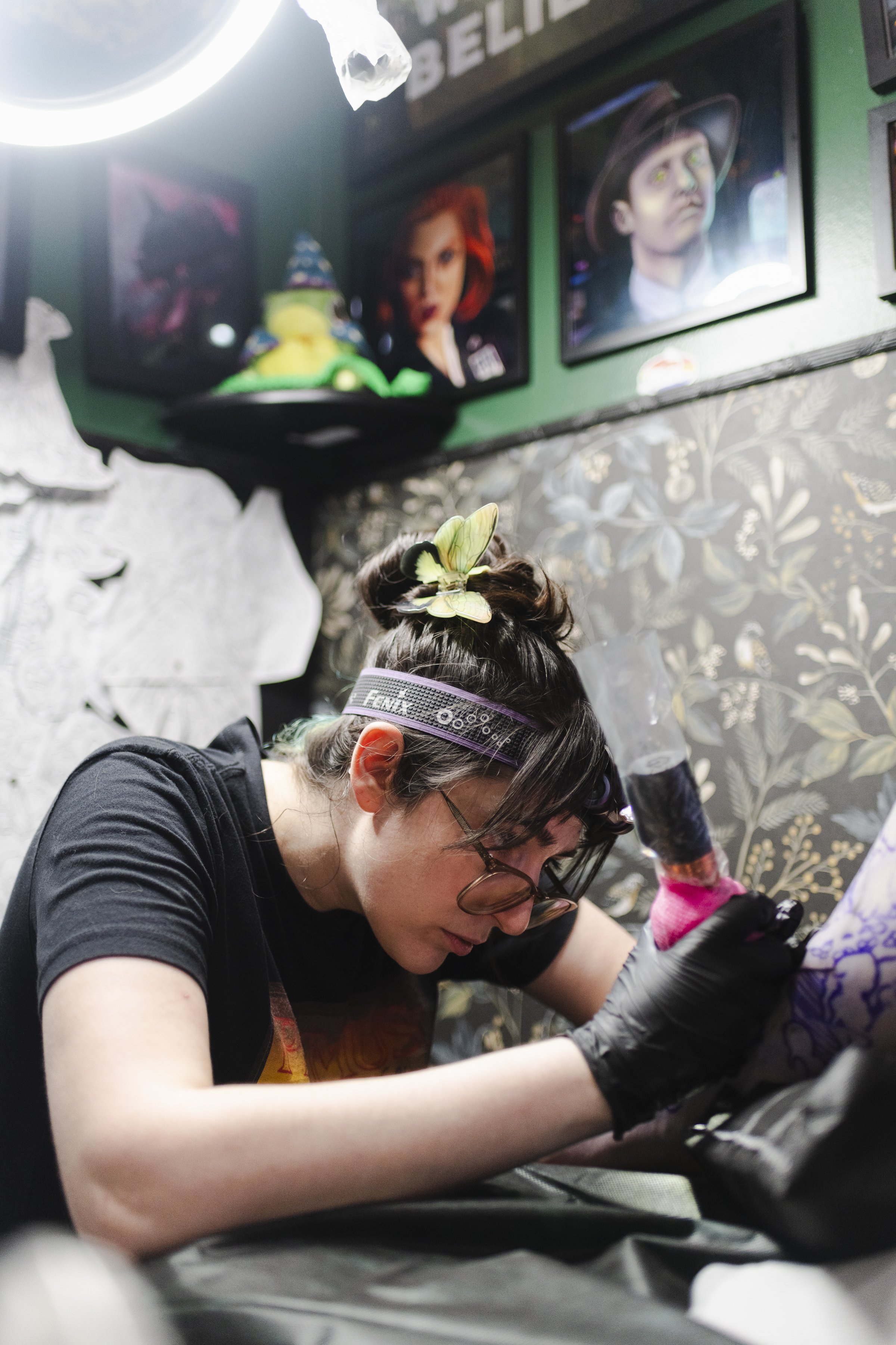 A woman with dark hair wearing glasses and a butterfly hair clip is tattooing skin, possibly doing a tattoo or similar detailed work, with a dark tattooed arm and a colorful floral background.