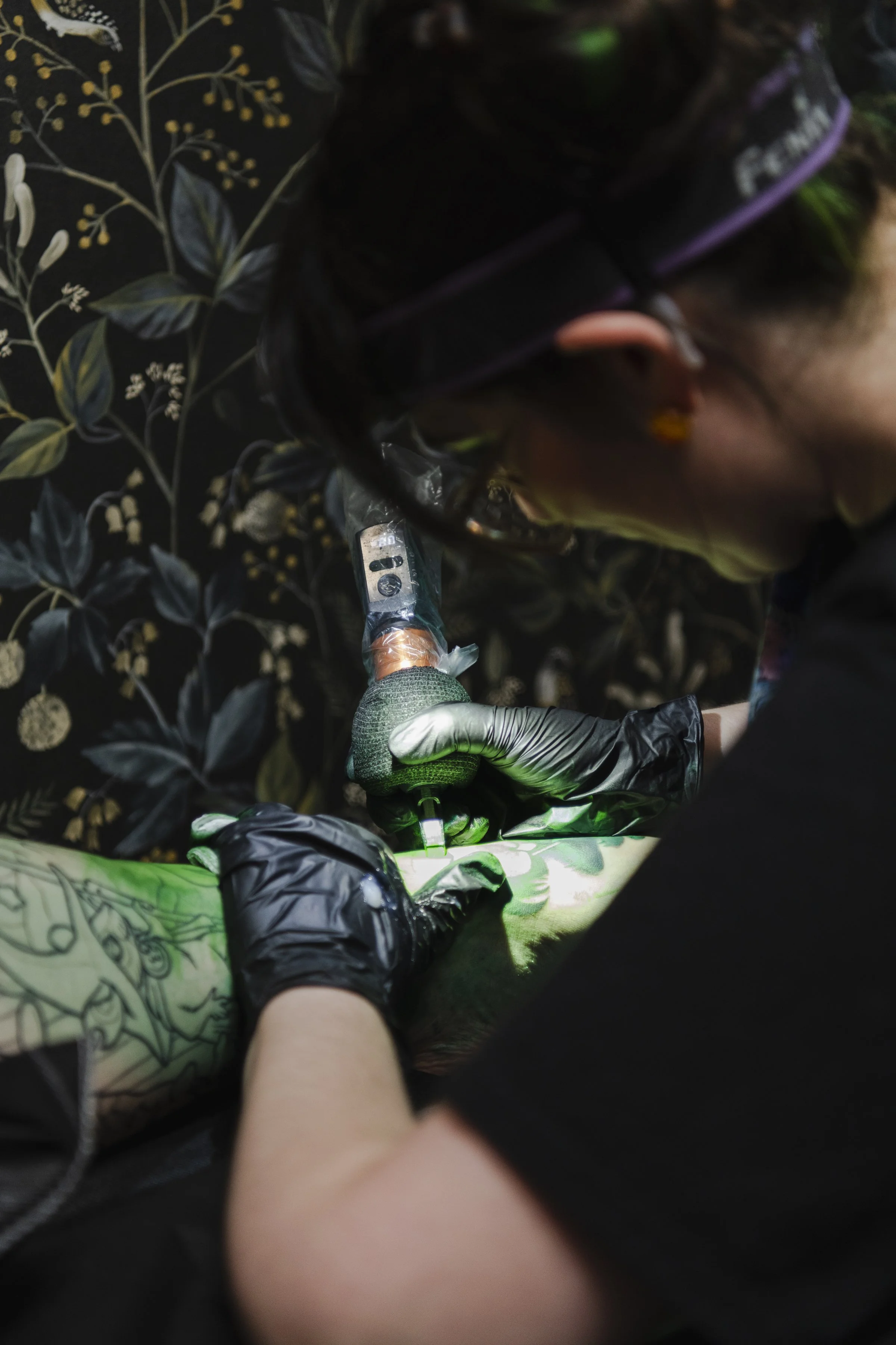 Person getting a tattoo with a tattoo gun while another person, likely the tattoo artist, is working on an arm that has a green tattoo design. The background is dark with a floral pattern.
