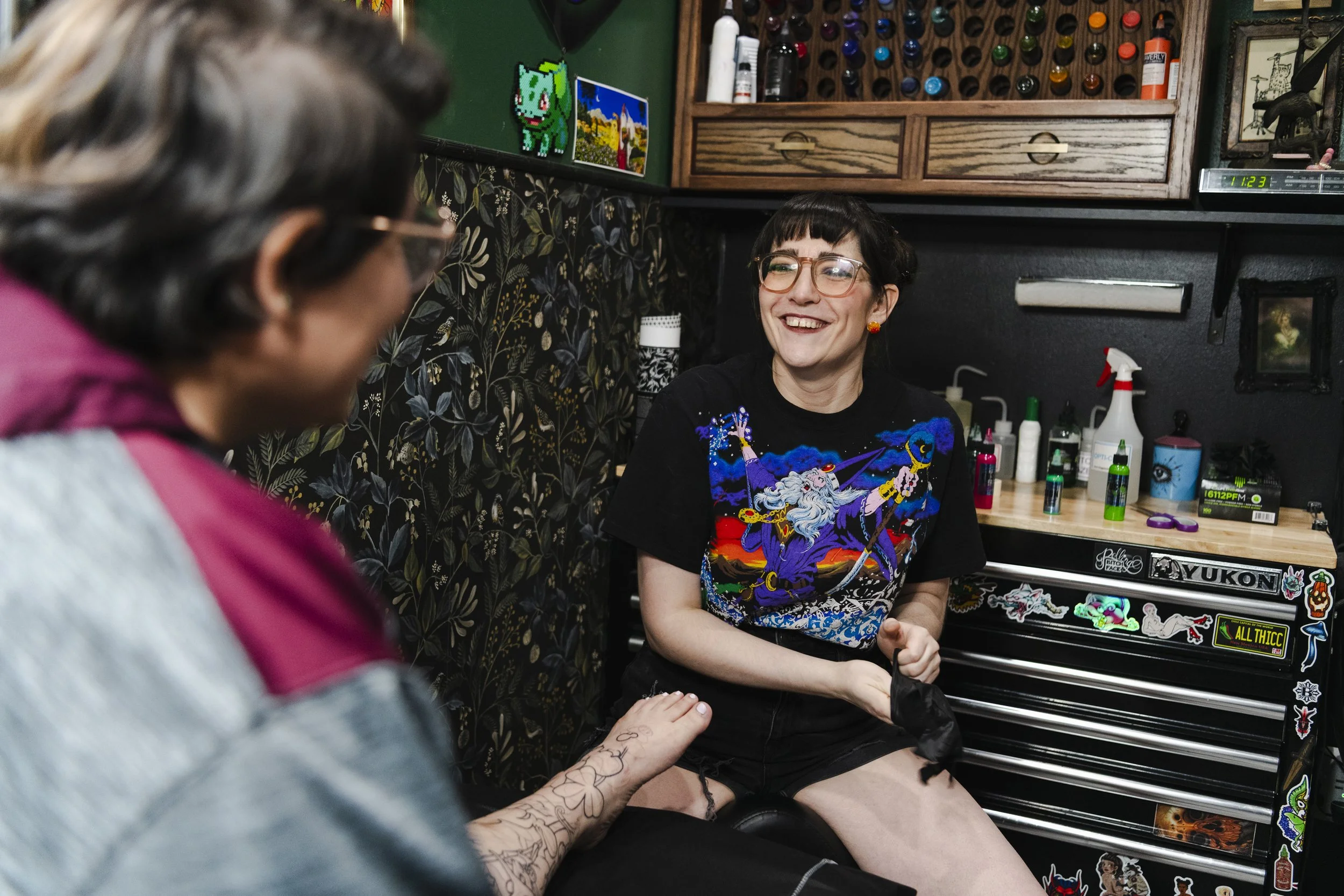 A smiling woman with dark hair, glasses, and earrings, sitting in a tattoo shop, having her tattoo inked by an artist with tattoos on his arm, in front of a tool chest with stickers.