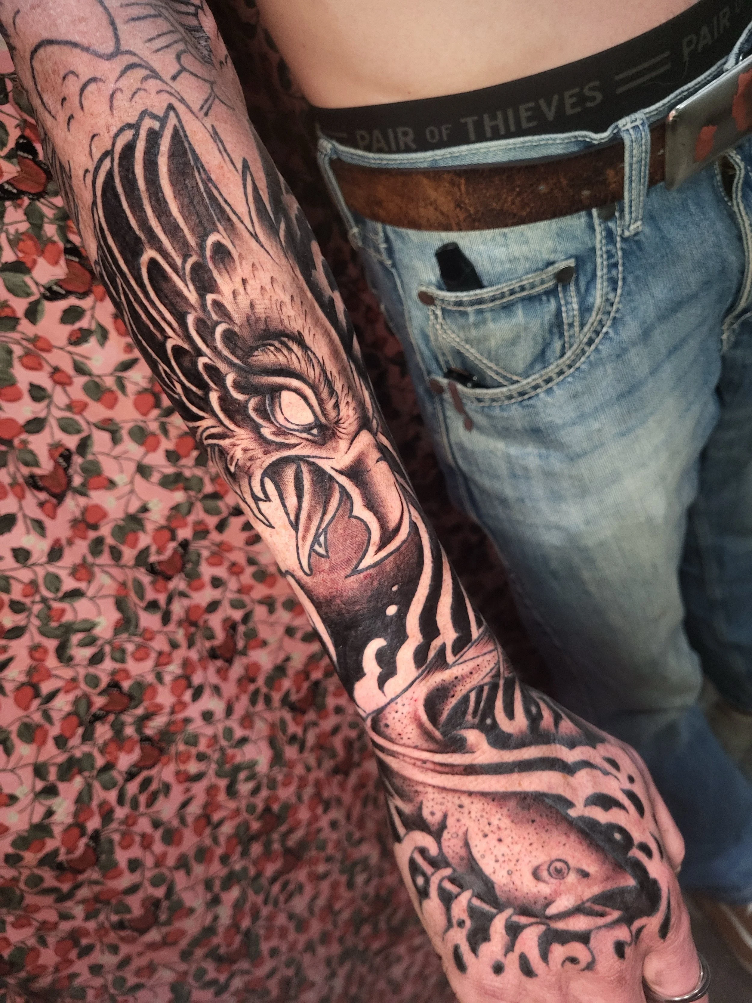 A person's arm with a detailed black and gray tattoo sleeve featuring a wolf, various abstract patterns, and some fine shading. The person is wearing a floral patterned shirt and has their arm resting on a pink and green patterned surface.