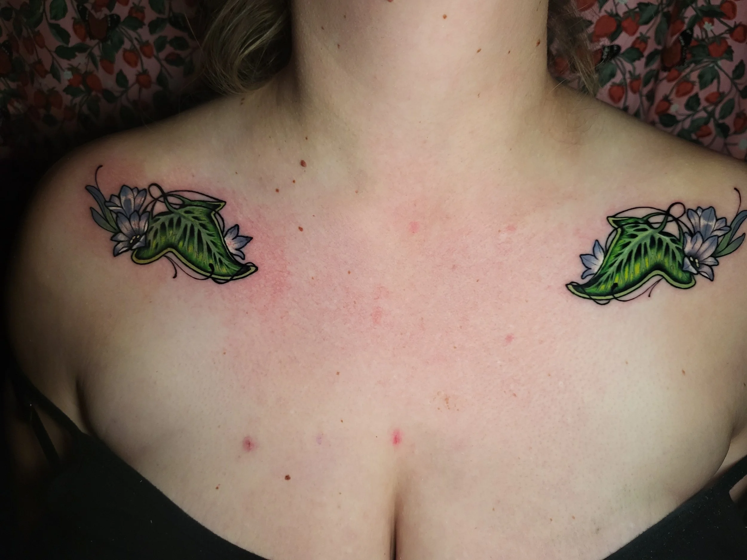 Close-up of a person's upper chest and shoulder area featuring tattoos of large green leaves and purple flowers.