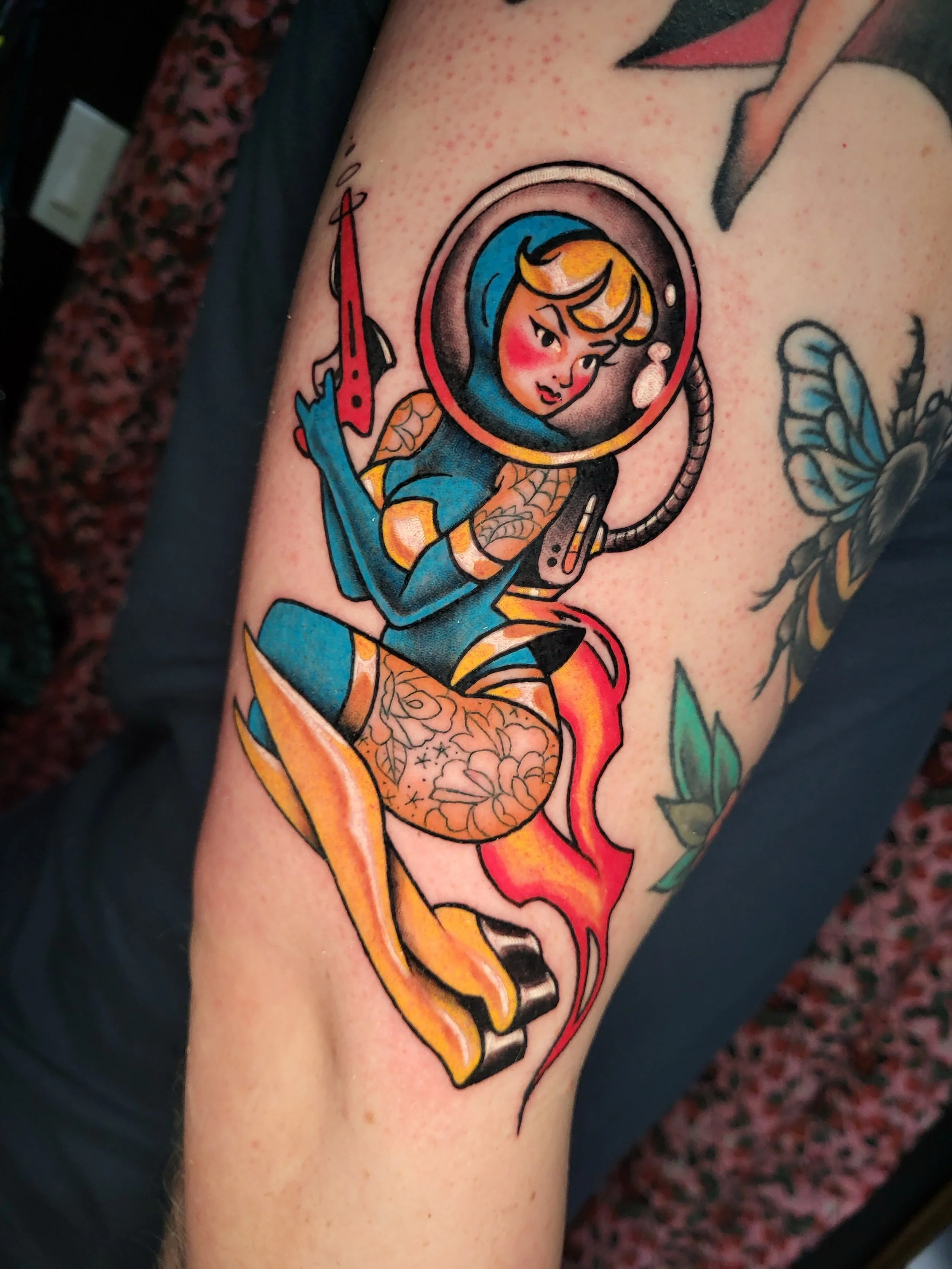 Tattoo of a cartoon girl in an astronaut suit with a fish tail, floating in space with a rocket and stars in the background.