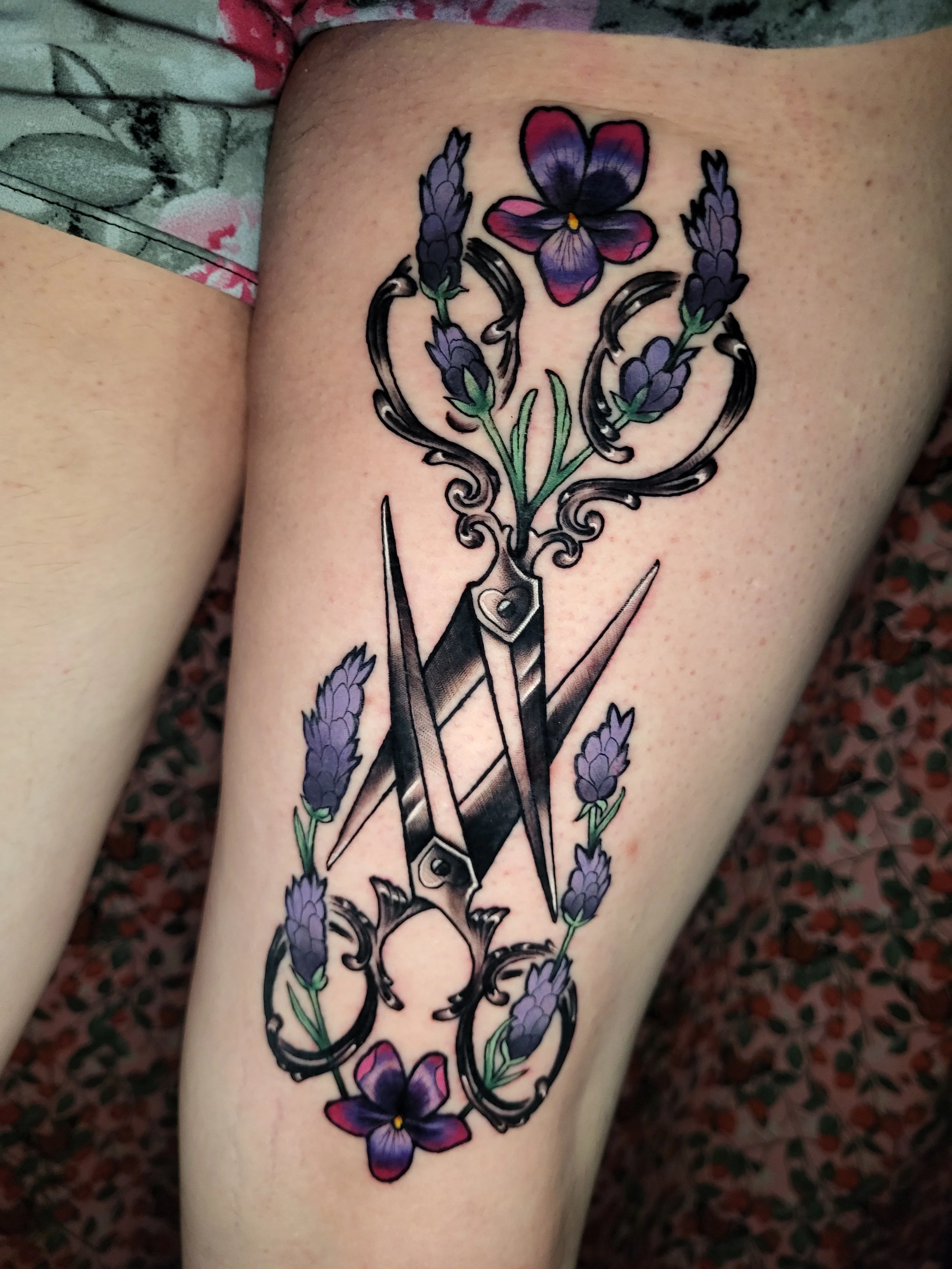 Tattoo of purple and pink pansy flowers arranged vertically along the forearm, intertwined with a decorative black scroll design and a central black dagger.