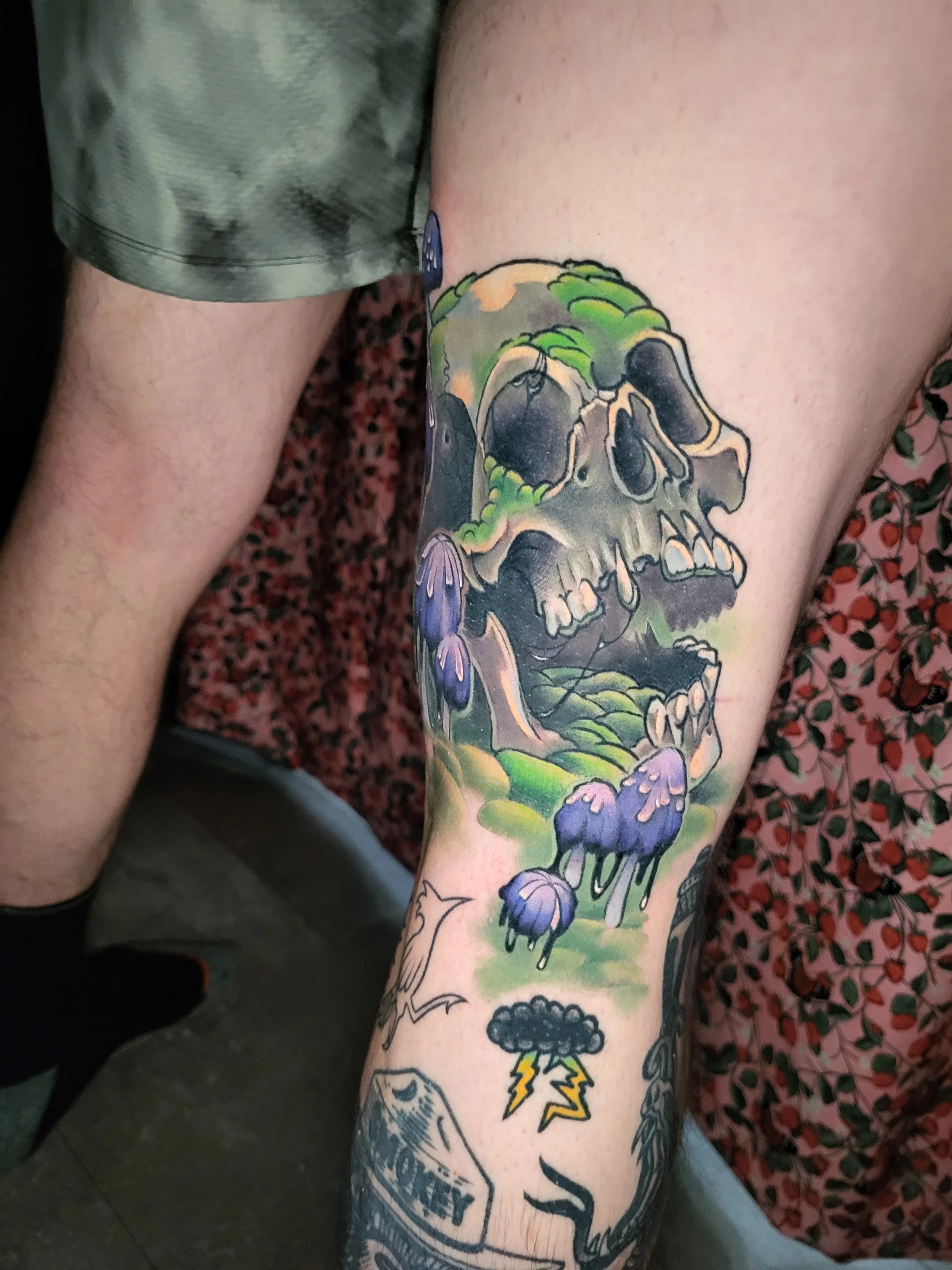 Tattoo of a skull with green leaves and purple flowers on a person's arm.