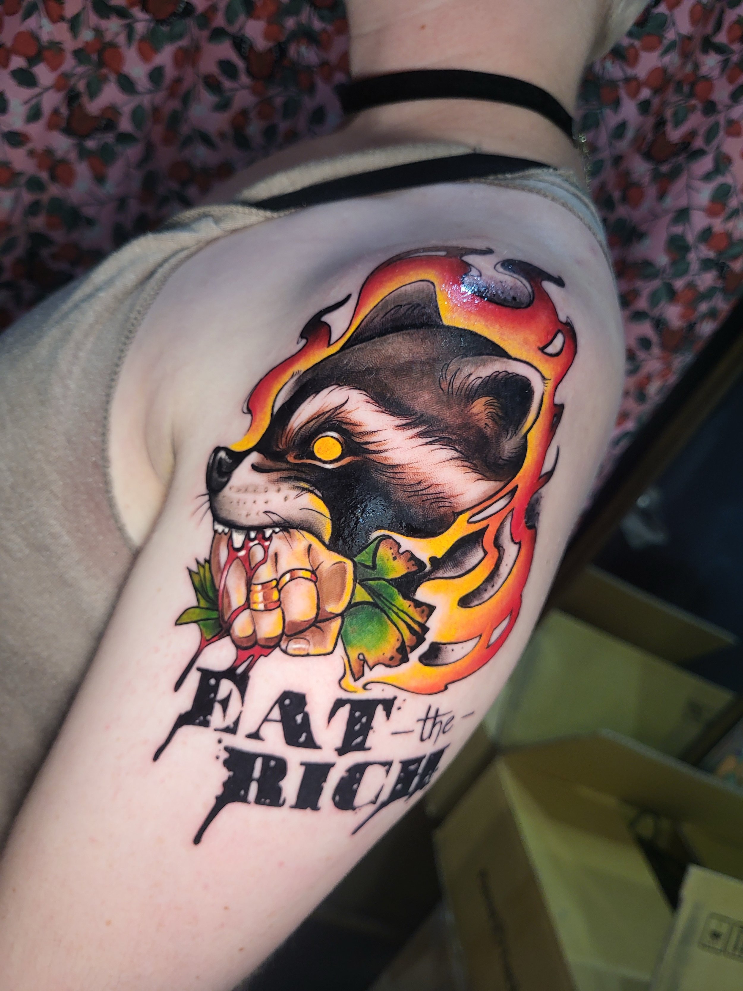 A colorful tattoo of a roaring lion with a fiery mane, a yellow eye, and a skull with flowers underneath on a person's arm.