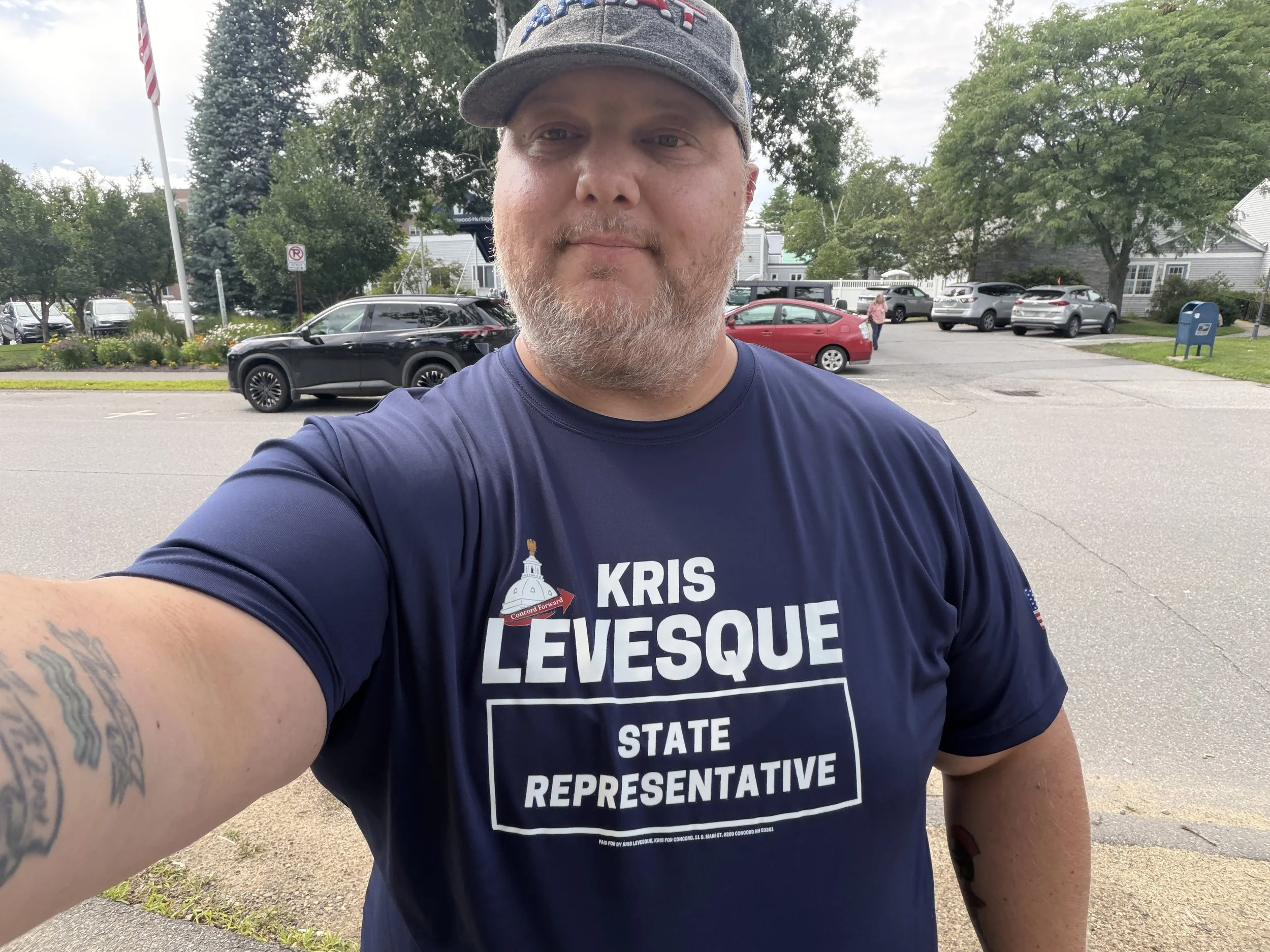 Kris Levesque in a campaign T-shirt reading "Kris Levesque, State Representative," outdoors