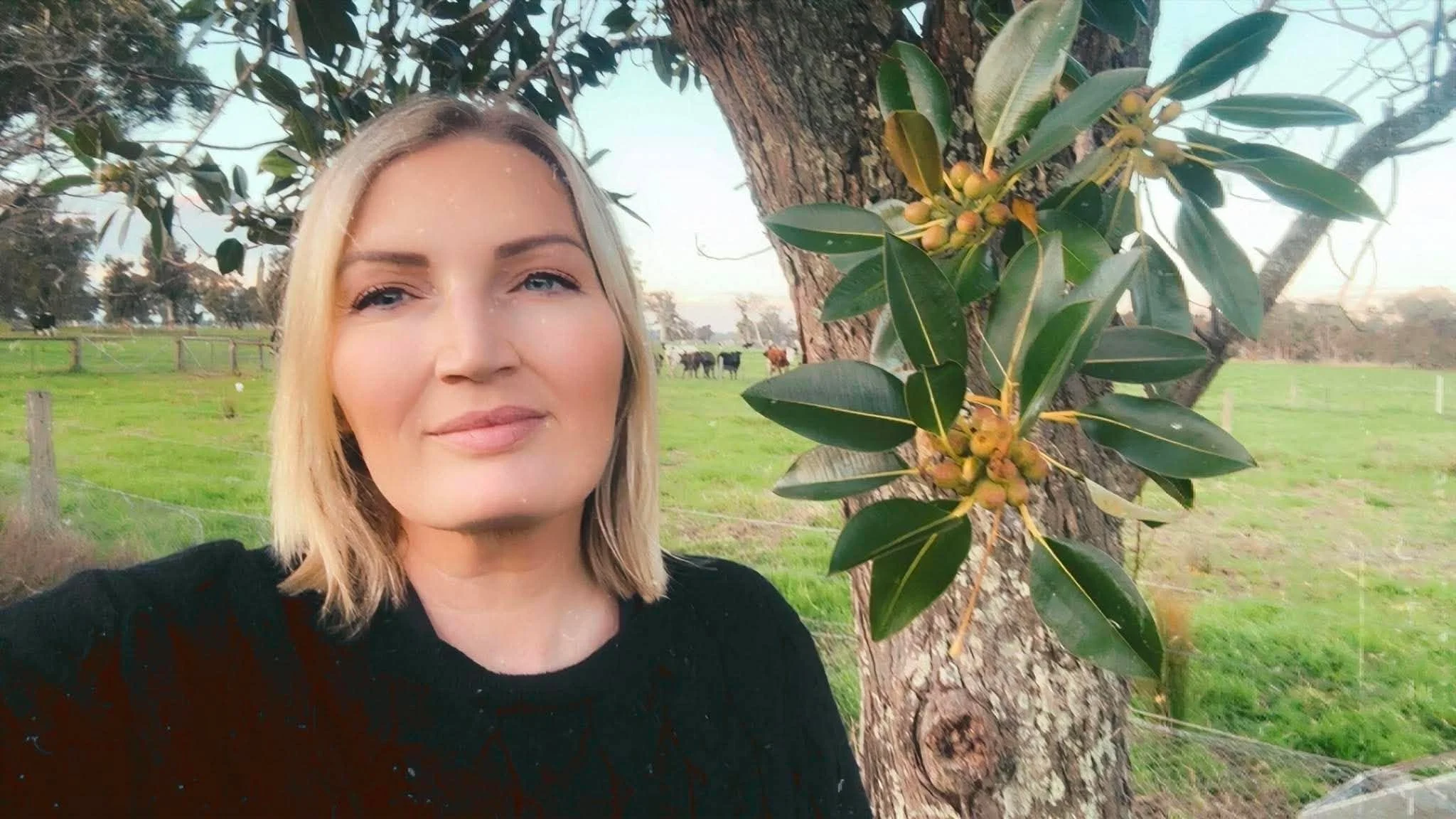 A woman with blonde hair and blue eyes taking a selfie outdoors next to a tree with green leaves and small yellow-green berries, in a rural setting with a grassy field and cows in the background.