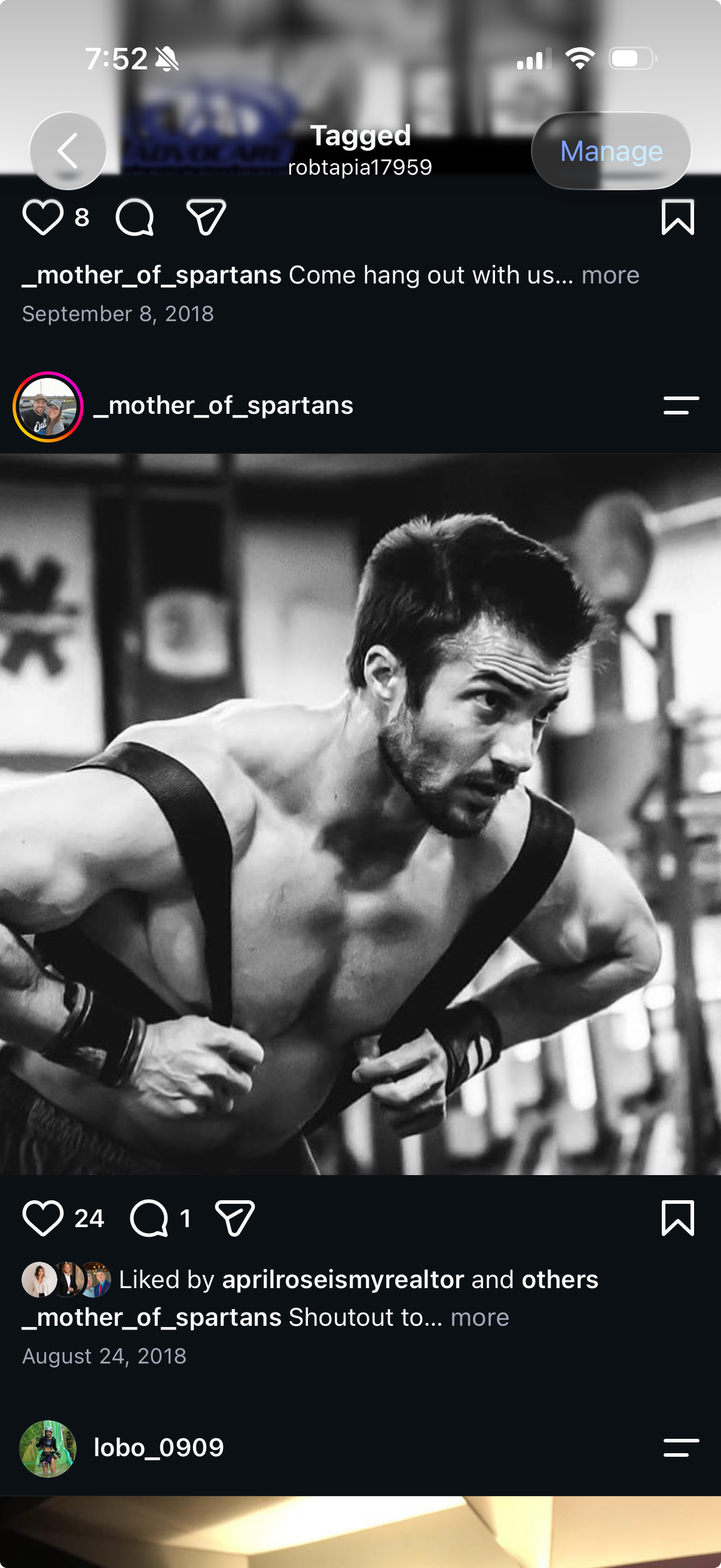A shirtless man with a beard and dark hair in a gym, leaning forward with hands on his hips, wearing wristbands and a tank top strap off, focused while posing for a photo in black and white.