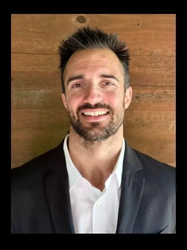 A smiling man with dark hair and a beard, dressed in a black suit and white shirt, posed in front of a wooden background.