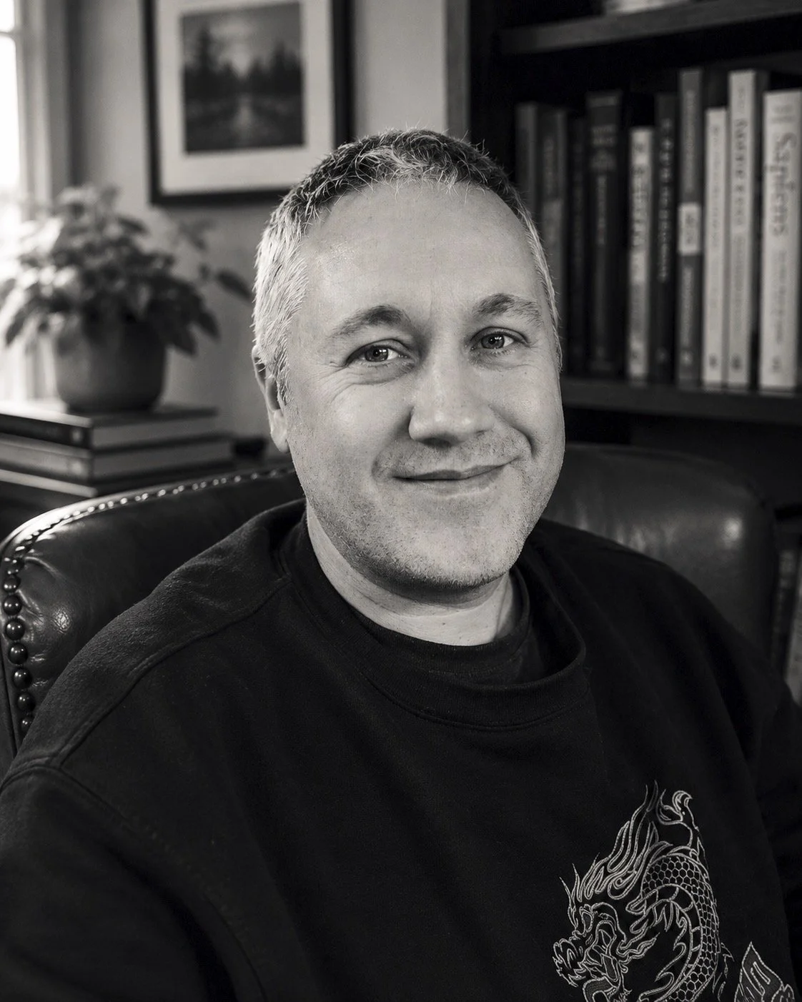 A black and white photo of a man with short hair, smiling while sitting in a leather chair in a room with bookshelves and a framed picture in the background.