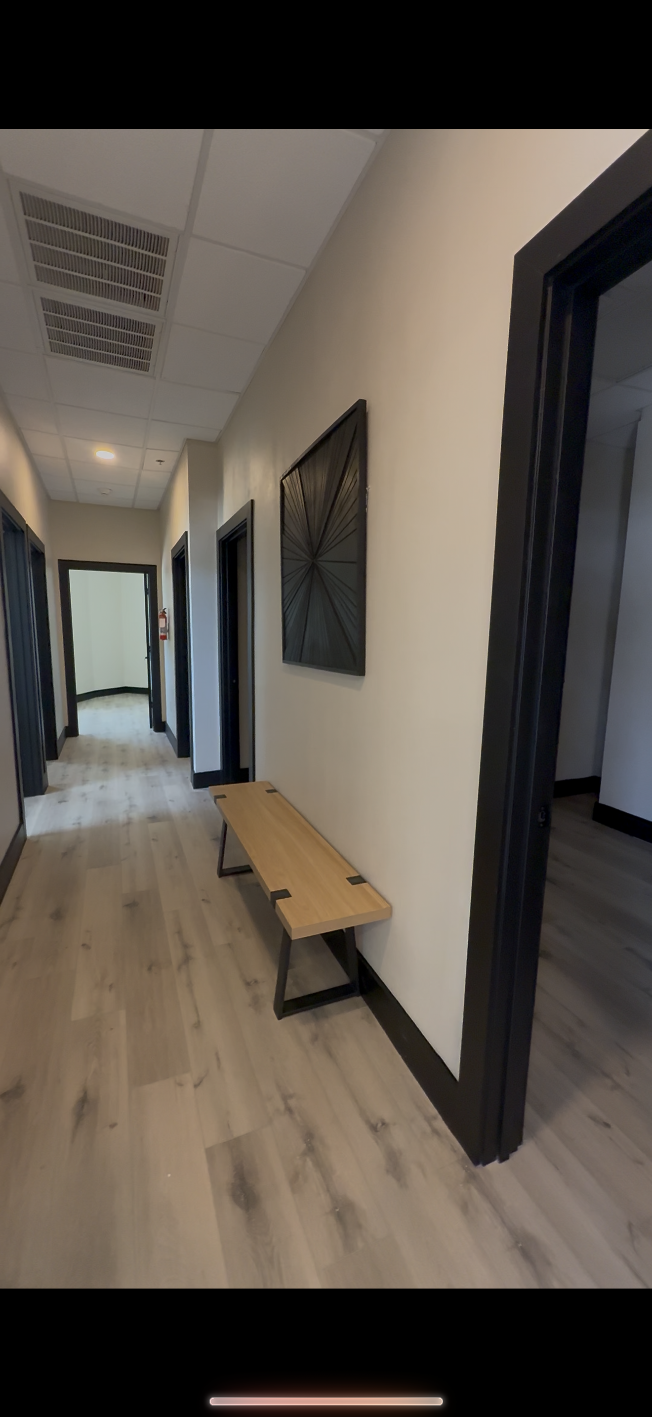 Hallway in an office building with wooden floor, black door frames, artwork on the wall, and a wooden bench.