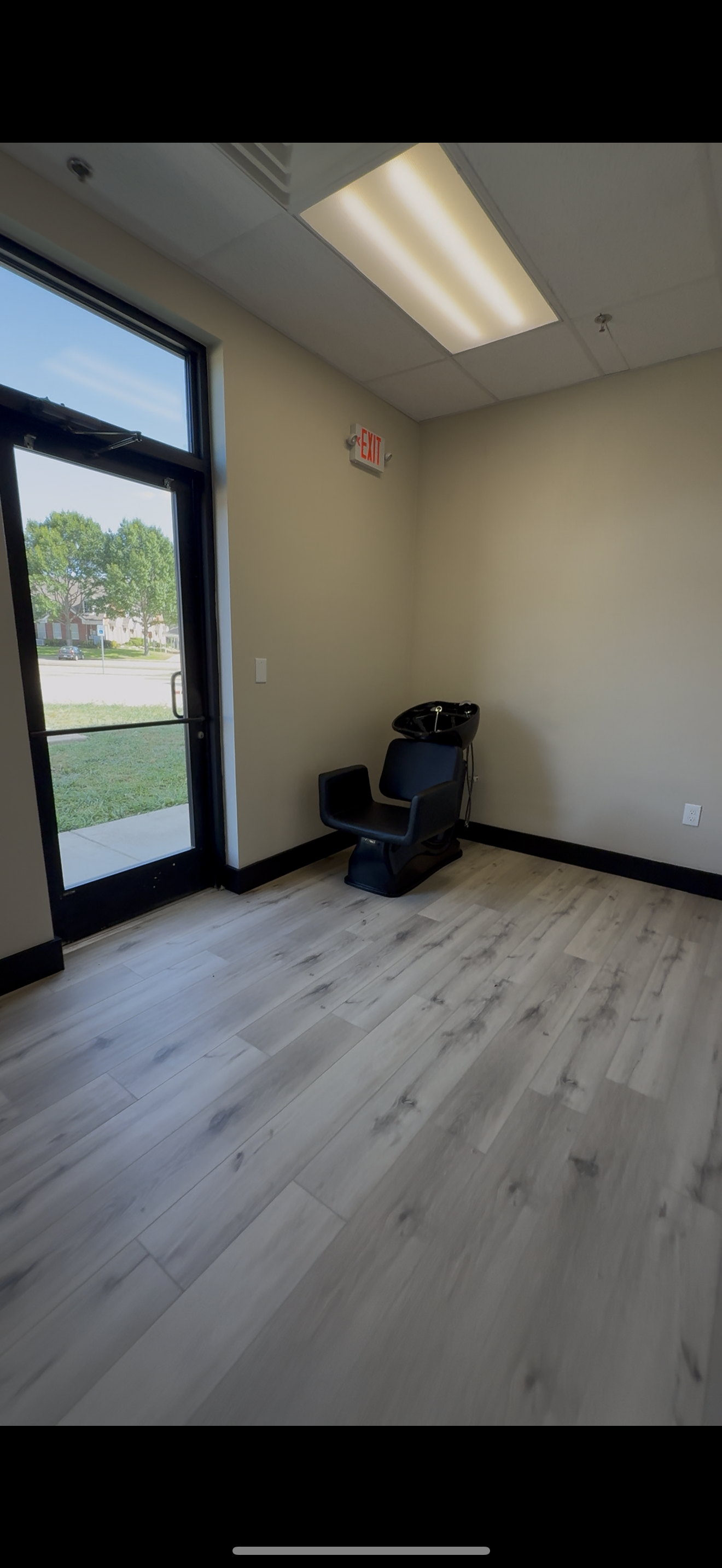 Empty hair salon corner with a black salon chair, a basin for washing hair, and an exit door with a glass window showing trees and houses outside.