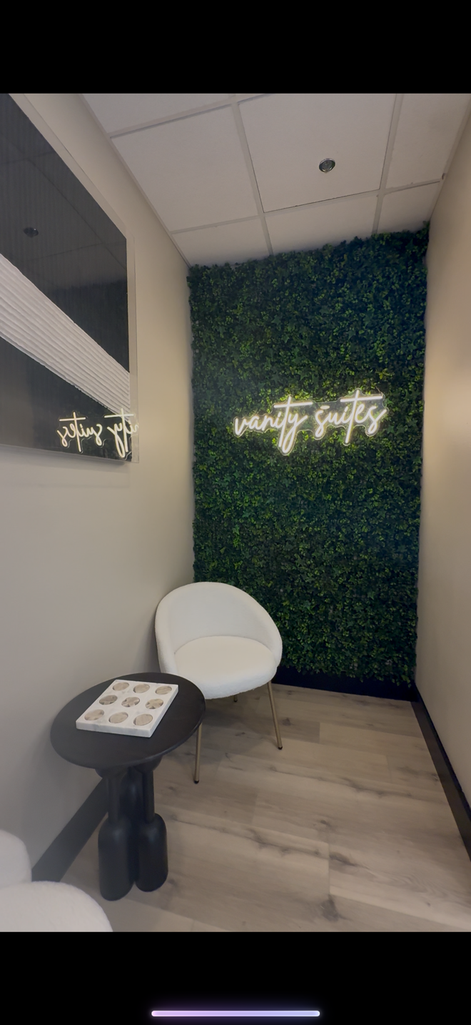 Cozy waiting area with a green artificial plant wall, a neon sign reading 'vanity suites,' a round white chair, a small black side table, and a decorative tray on the table.