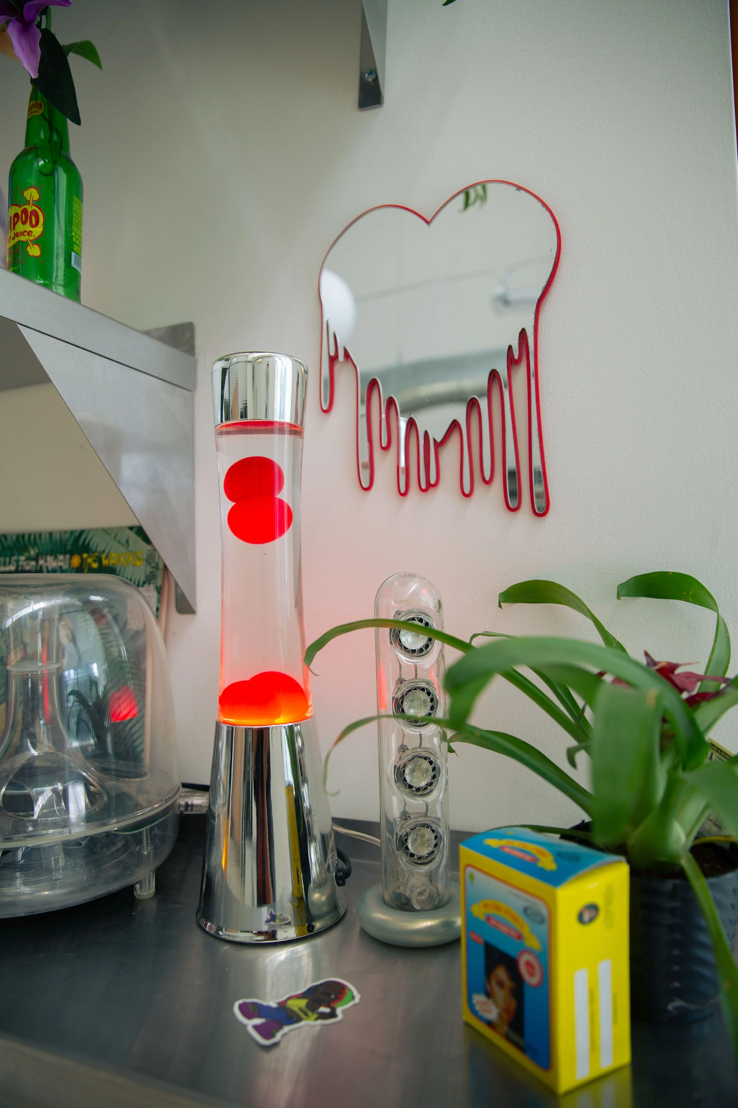 A vaporizer lamp with red liquid inside, a plant in a pot to the right, a yellow toy box, and some glassware on a metal surface. In the background, a mirror with red dripping design and a green plastic bottle on a shelf.