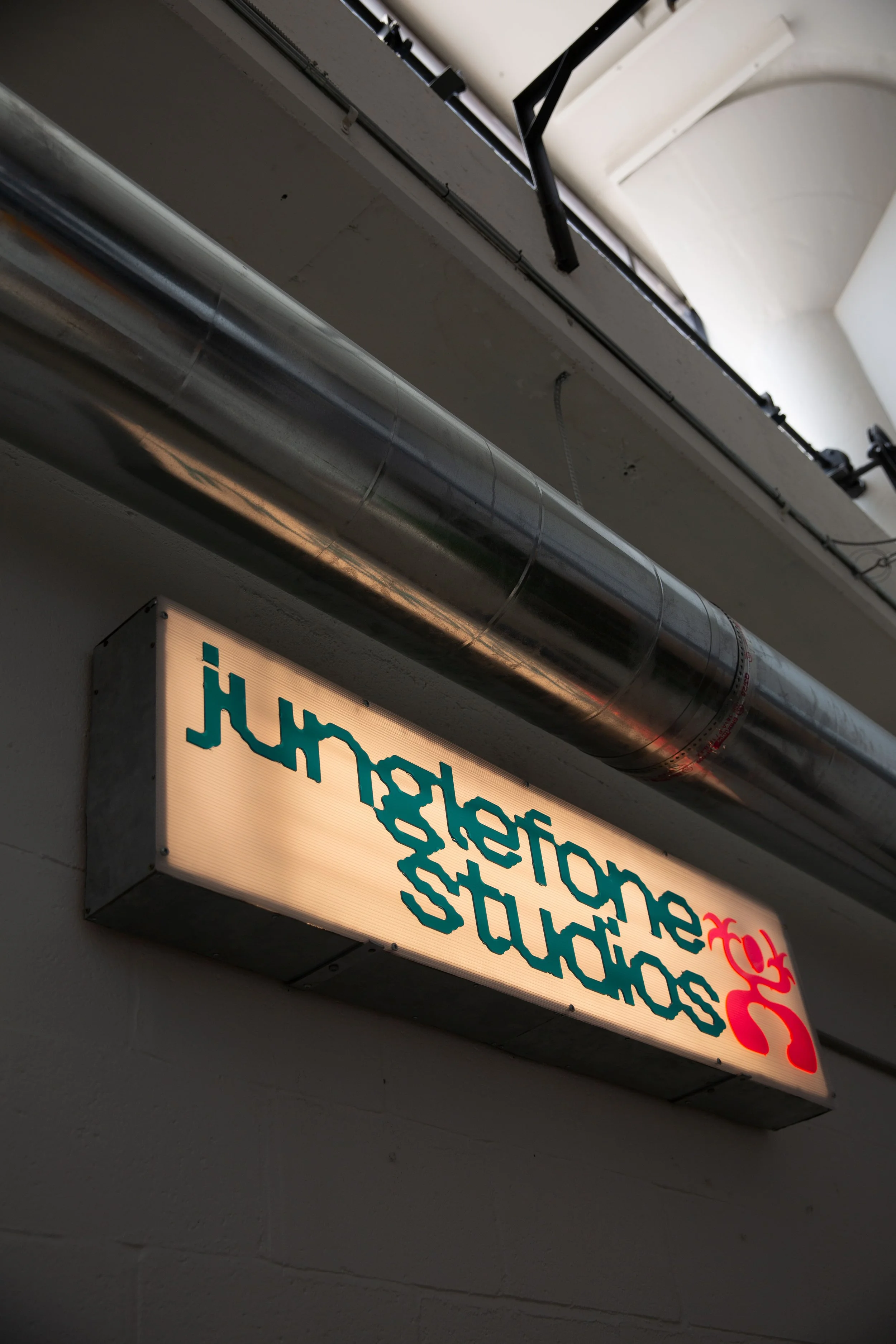 Close-up of an illuminated signboard displaying text in reverse, hanging on a wall near a metallic pipe and a ventilation duct.