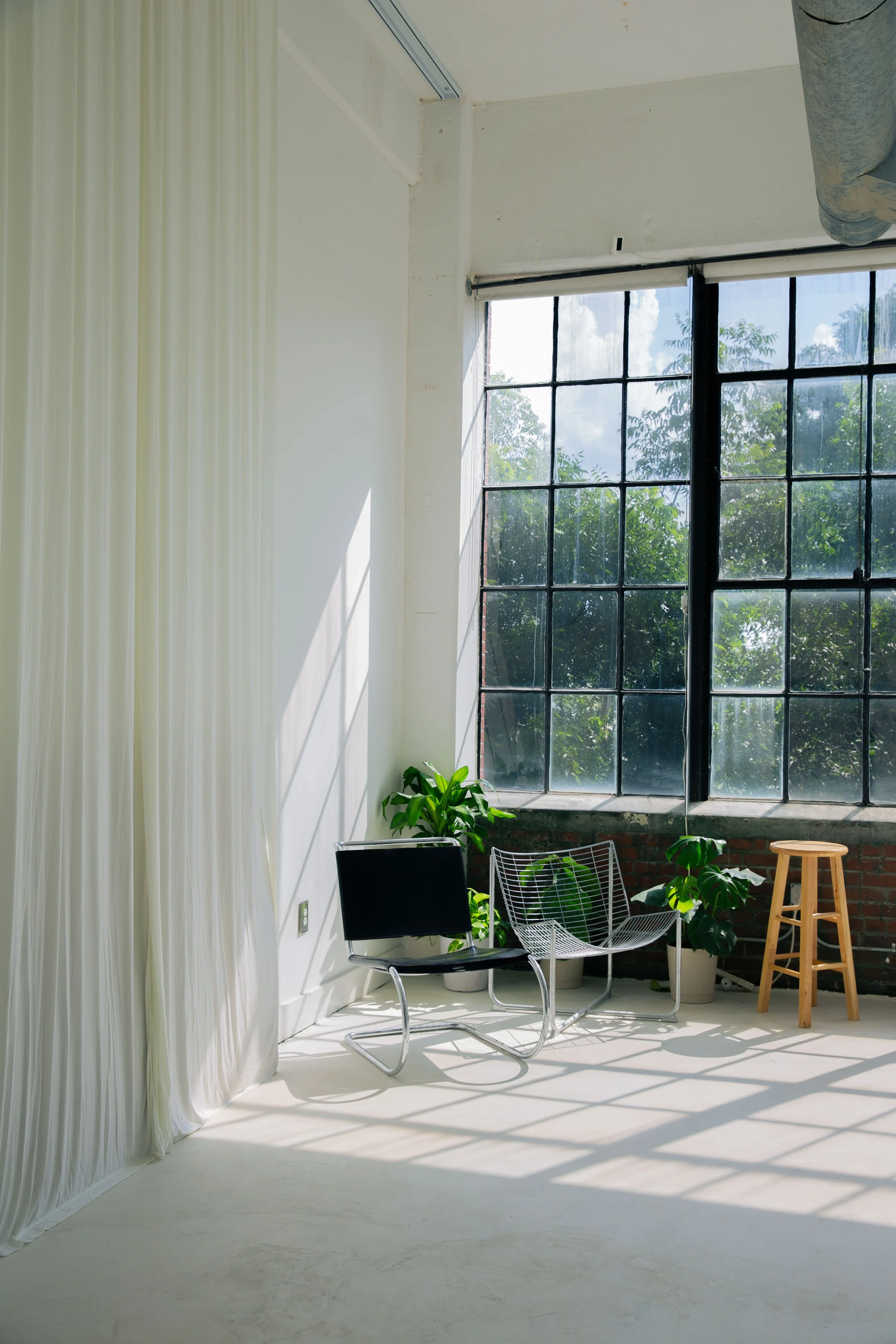 Interior of a bright room with large windows, white curtains on the left, black and wire chairs, plants, and a wooden stool.