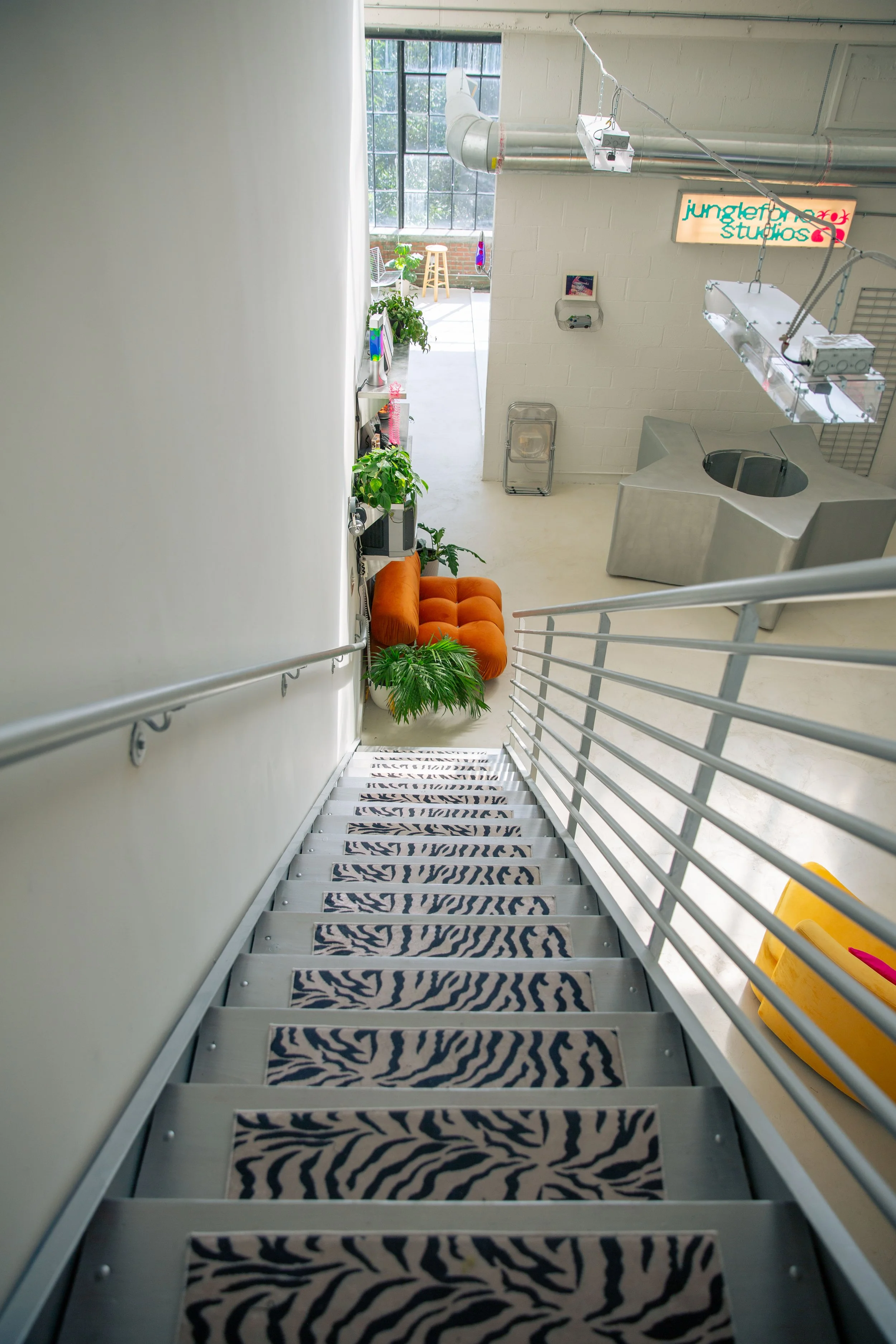 View from the top of a metal staircase with zebra-patterned stair treads, leading down to a room with a neon sign that says 'jungerfox studios' and various furniture and plants near large windows.