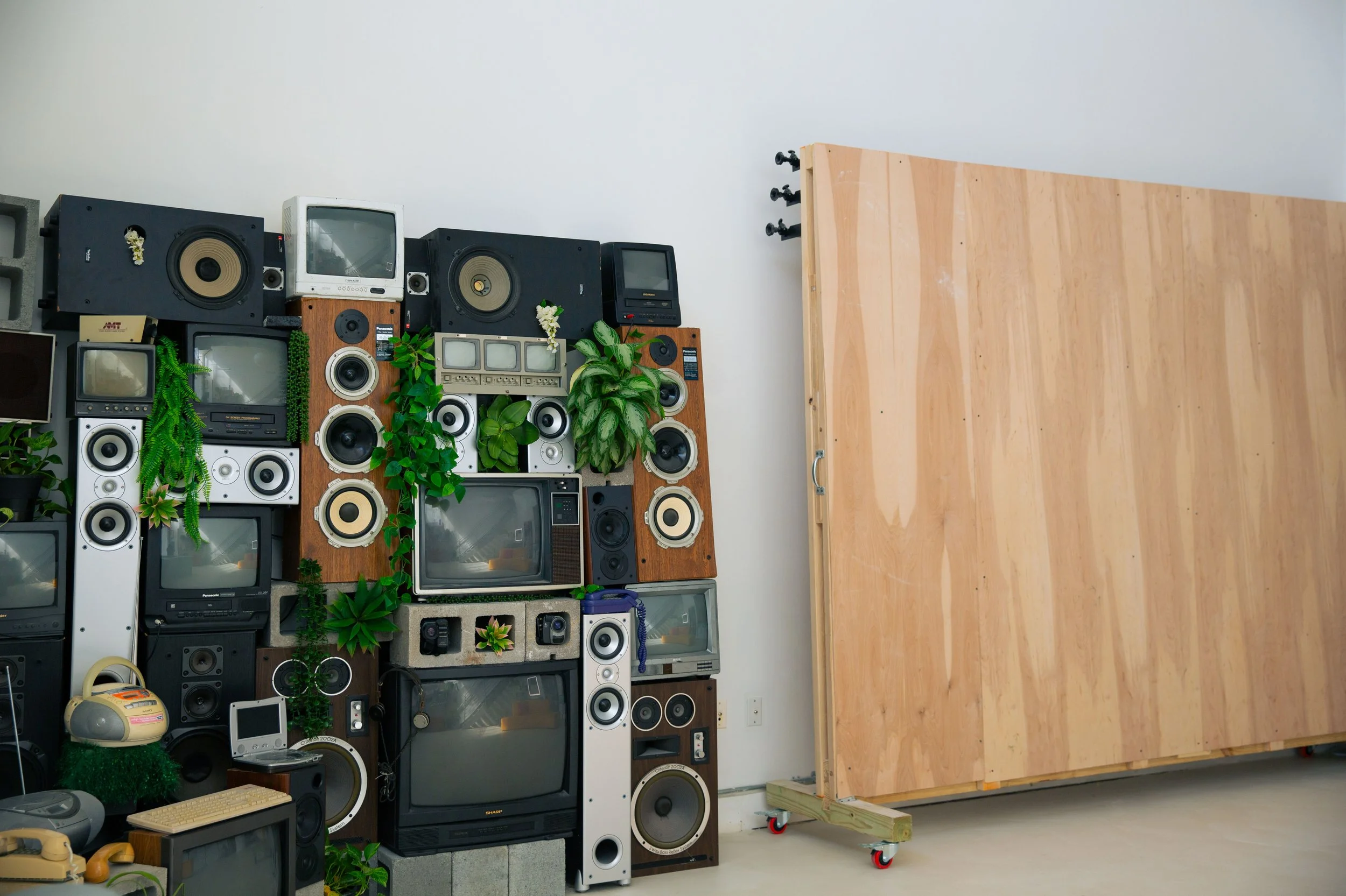 Collection of vintage televisions, radios, and speakers arranged against a white wall, with potted plants and a wooden moving cart nearby.
