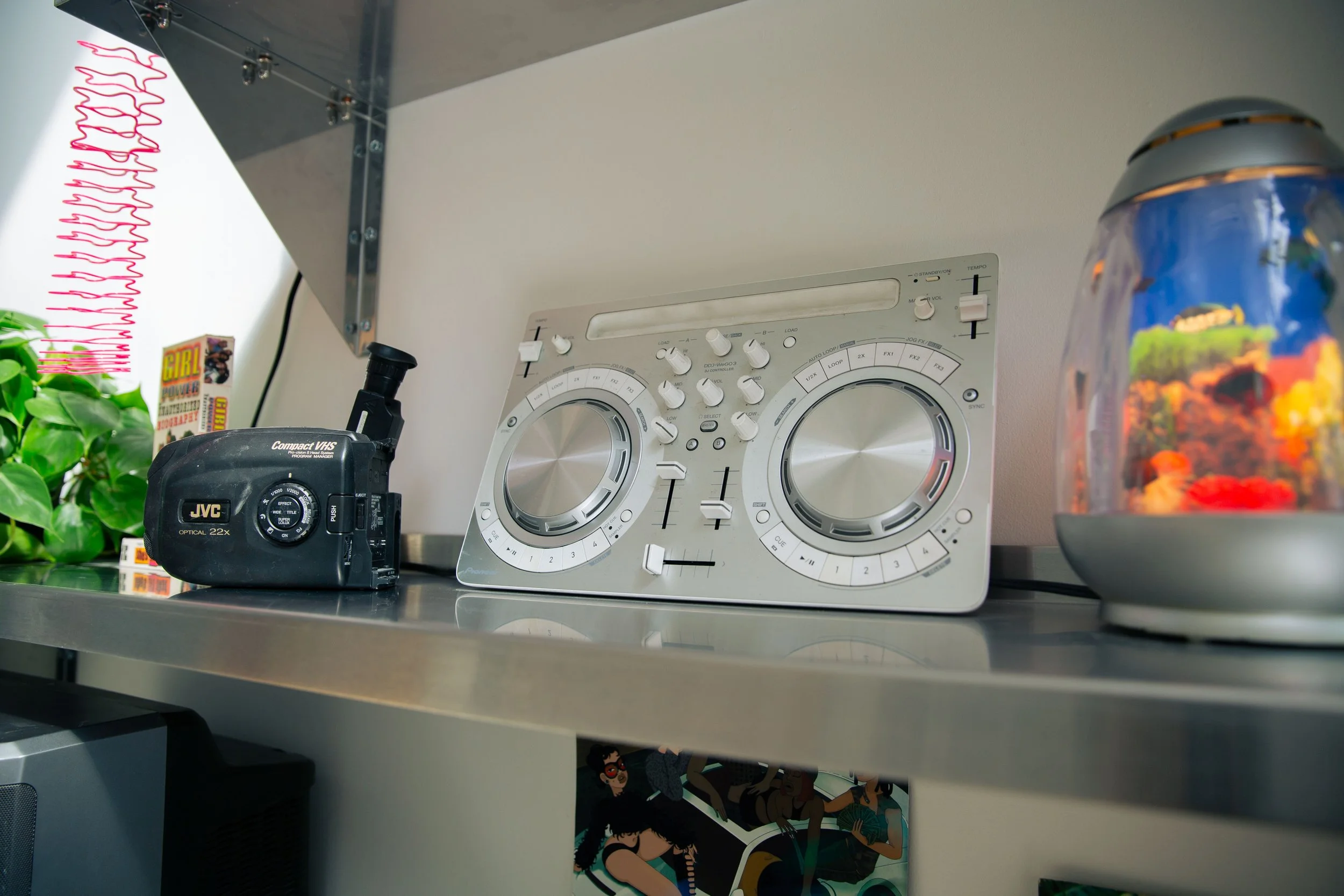 Shelf with a DJ mixing console, a vintage camera, some colorful artwork, plants, and a lava lamp.