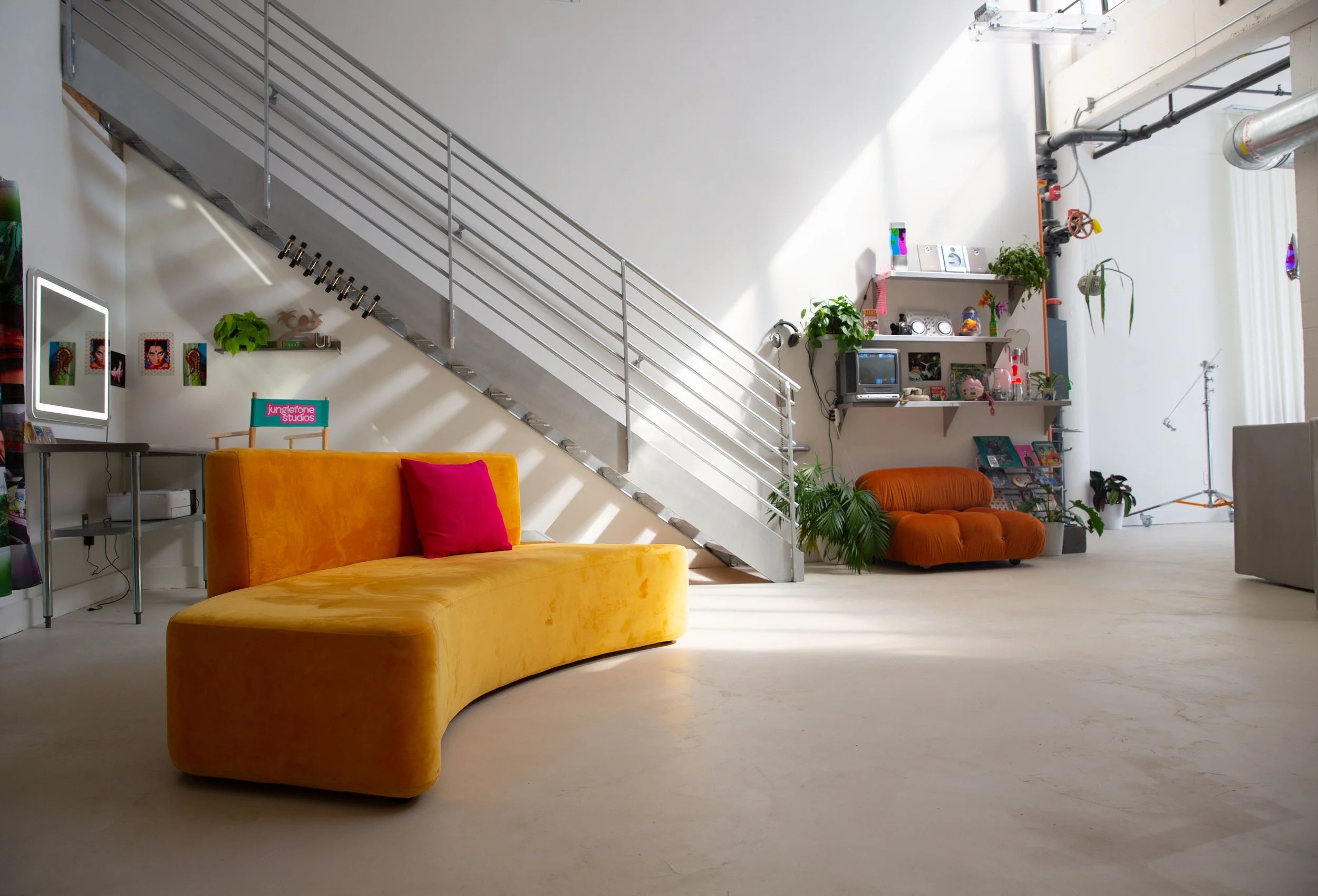 A bright, modern living room with a yellow velvet sofa with red pillows, a white wall with art and plants, a metal staircase, and various shelves with electronics, plants, and decorations. Natural light illuminates the space.
