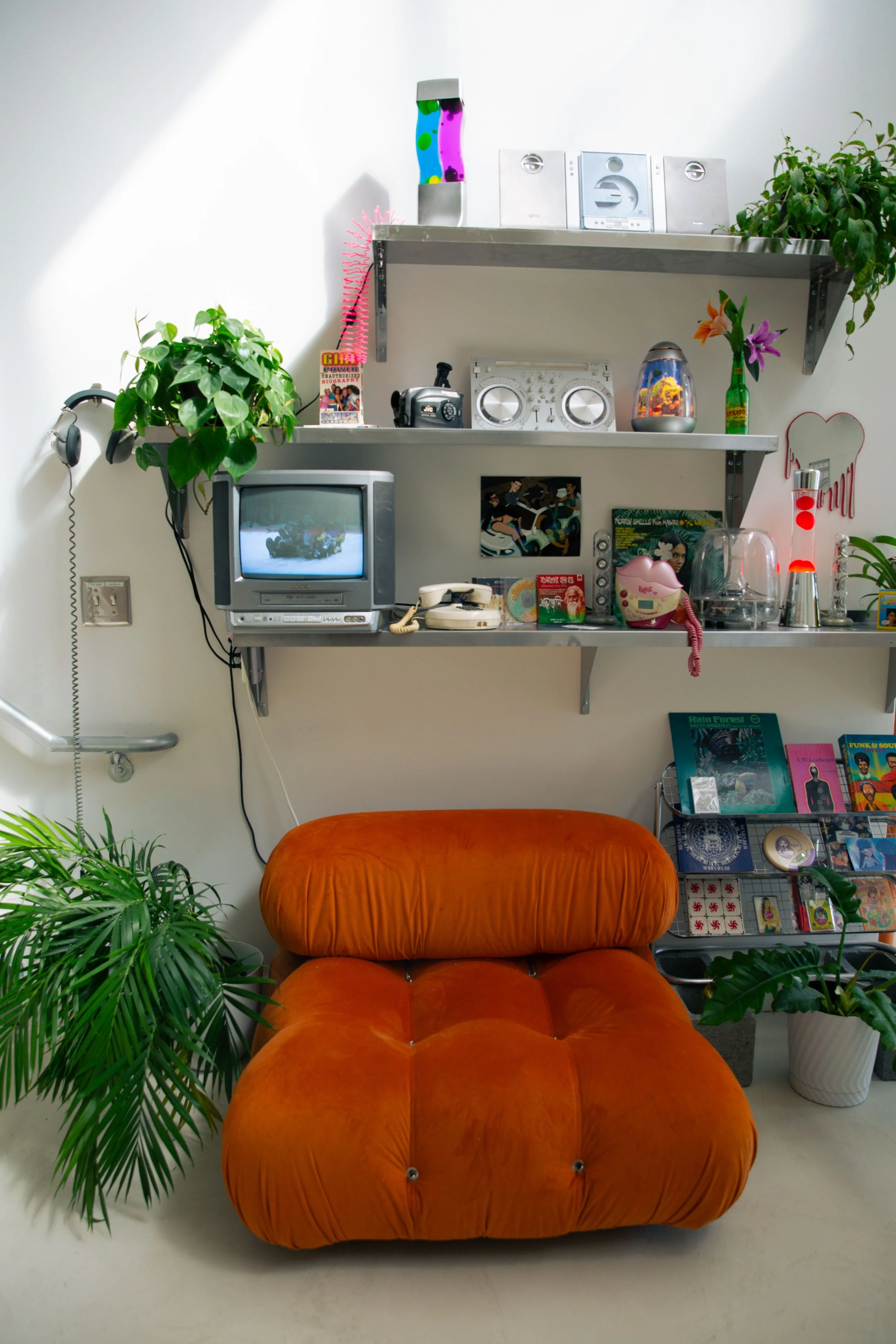 A room with a bright orange plush chair centered in front, green plants on either side, and a wall with shelves holding electronic devices, music equipment, and decorative items. A vintage TV, books, and magazines are also visible.