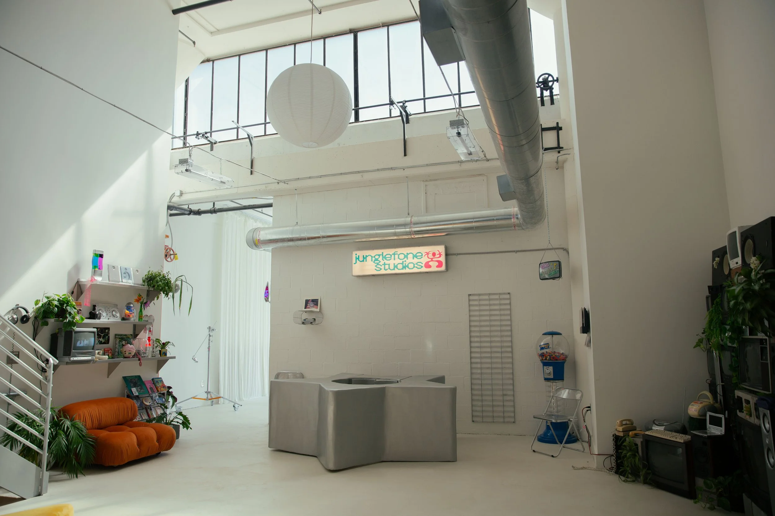 Interior of a spacious loft-style room with white walls and high ceilings, large windows, a leather sofa, plants, a shelf with various items, and visible ventilation ducts overhead.