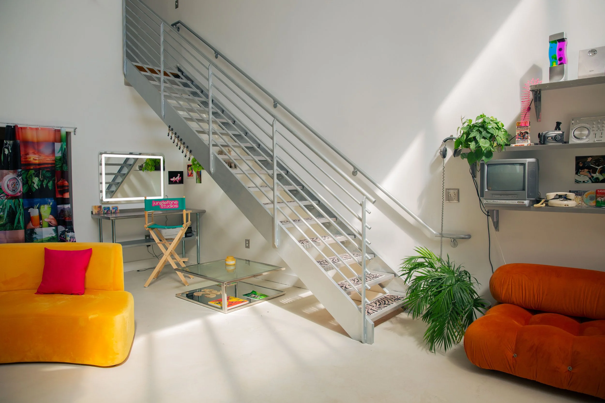 A cozy living room with a metal staircase leading to an upper floor, decorated with plants, colorful furniture, and vintage electronics, under natural light.