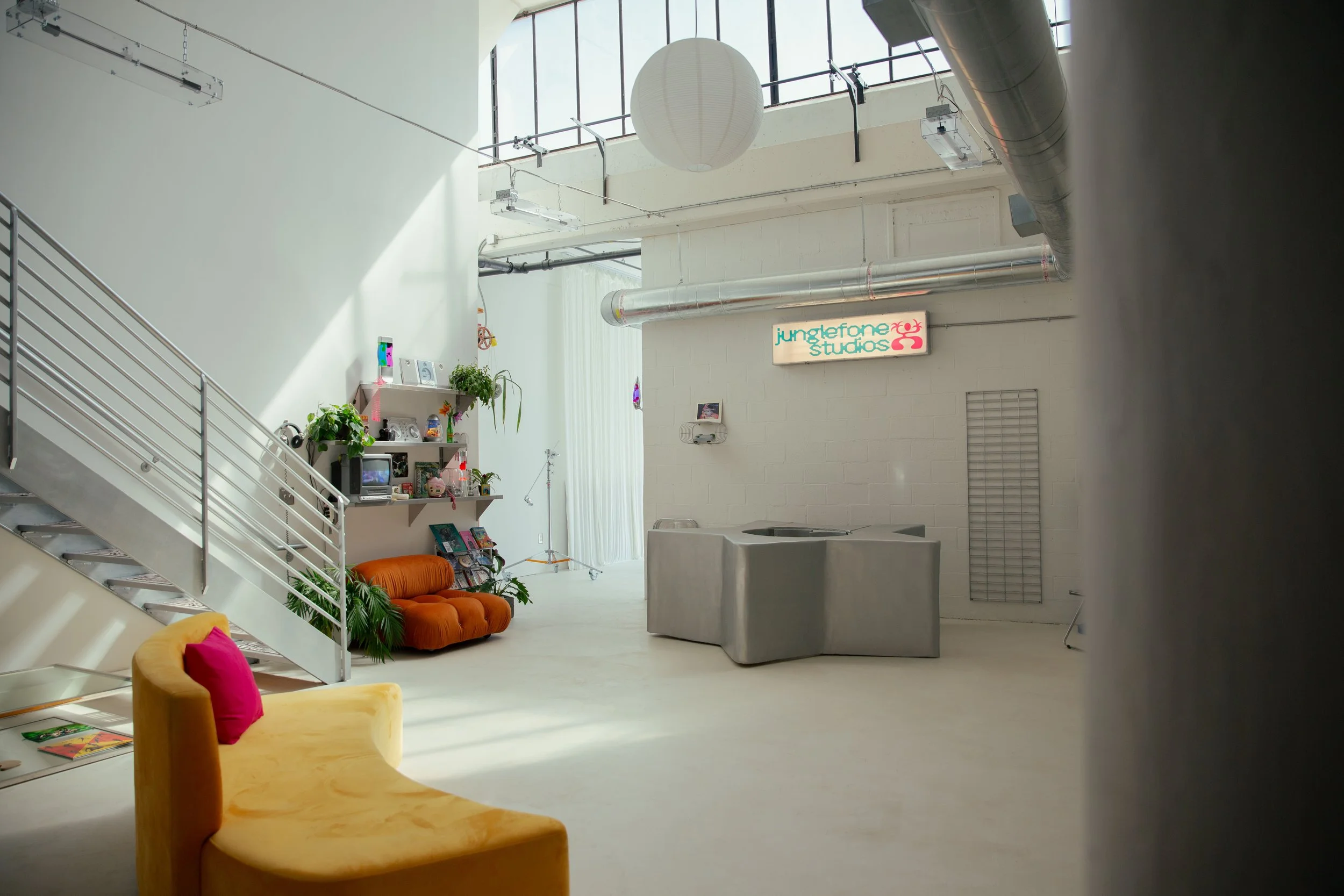 Interior of a modern spacious studio with white walls and large windows, featuring a yellow curved chair with pink pillows, a green couch, a set of shelves with plants and decorative items, a sign that reads "junglefone studios," a gray armchair, and exposed ductwork and lighting fixtures.