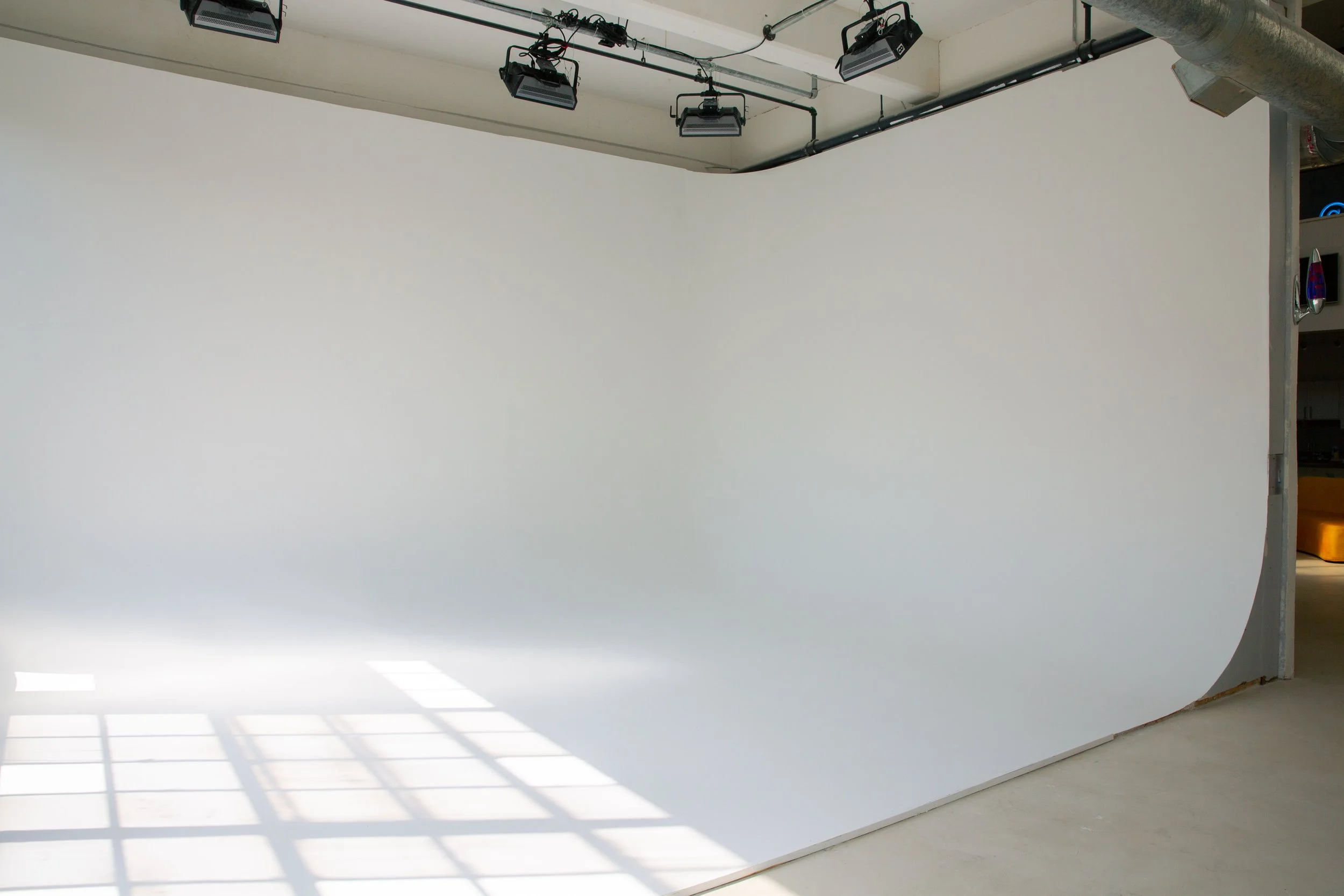A photography studio with a large white backdrop, three studio lights hanging from the ceiling, and sunlight casting a pattern of shadows on the floor. There are some objects visible on the right side, including a lava lamp and some other small items.