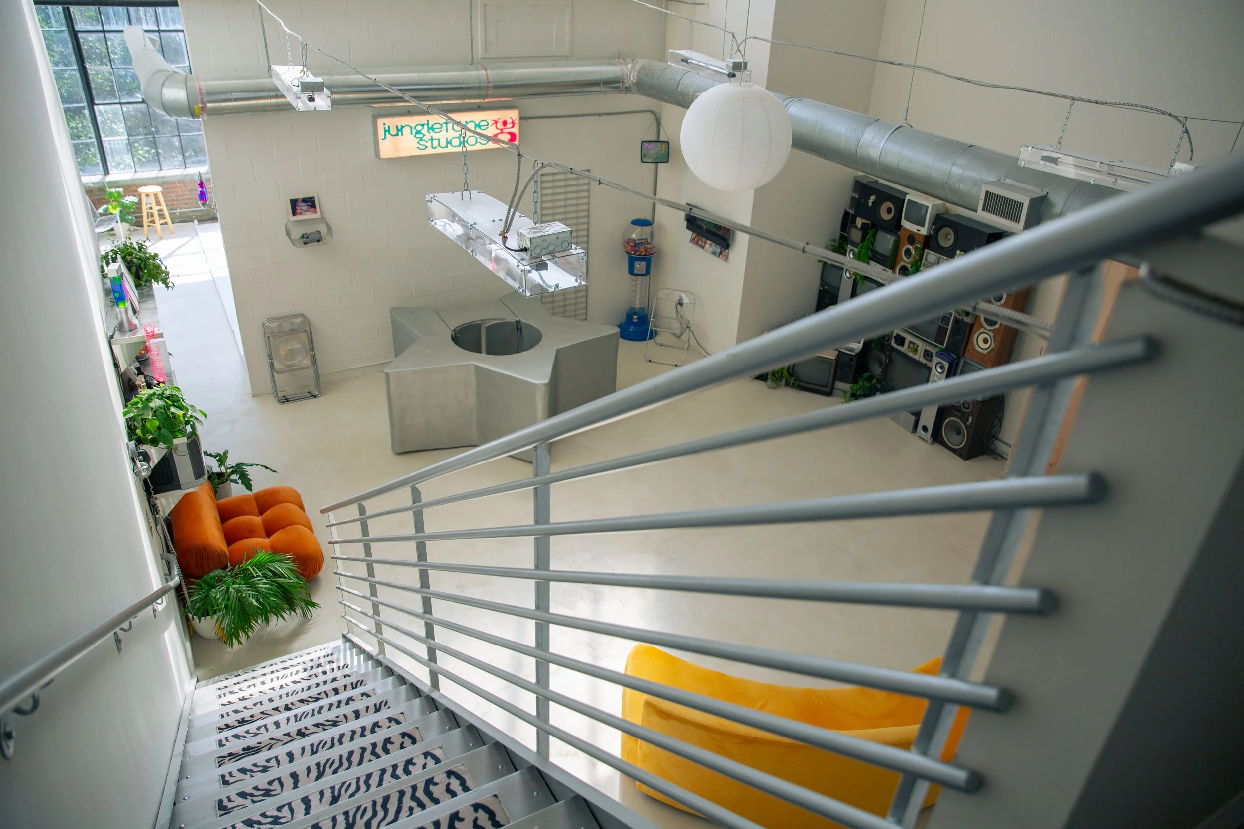 Interior of a modern loft apartment viewed from a staircase. The room features a neon sign reading 'jungle of the studio,' a large white paper lantern, a collection of speakers and audio equipment, a small television, and a kitchenette with plants, colorful chairs, and a window letting in natural light.